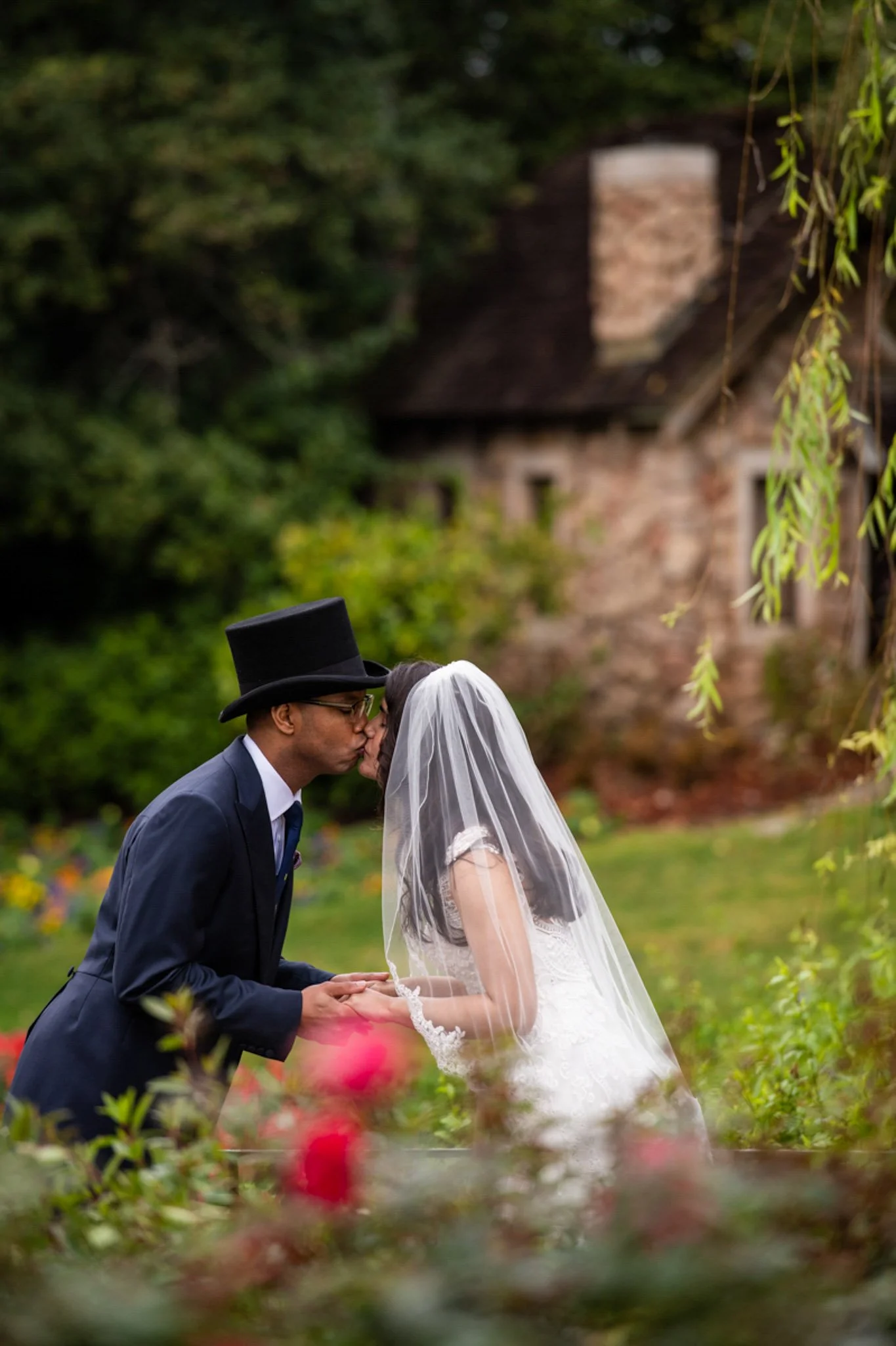 A couple dressed in wedding attire sharing a kiss outdoors with a rustic house in the background.