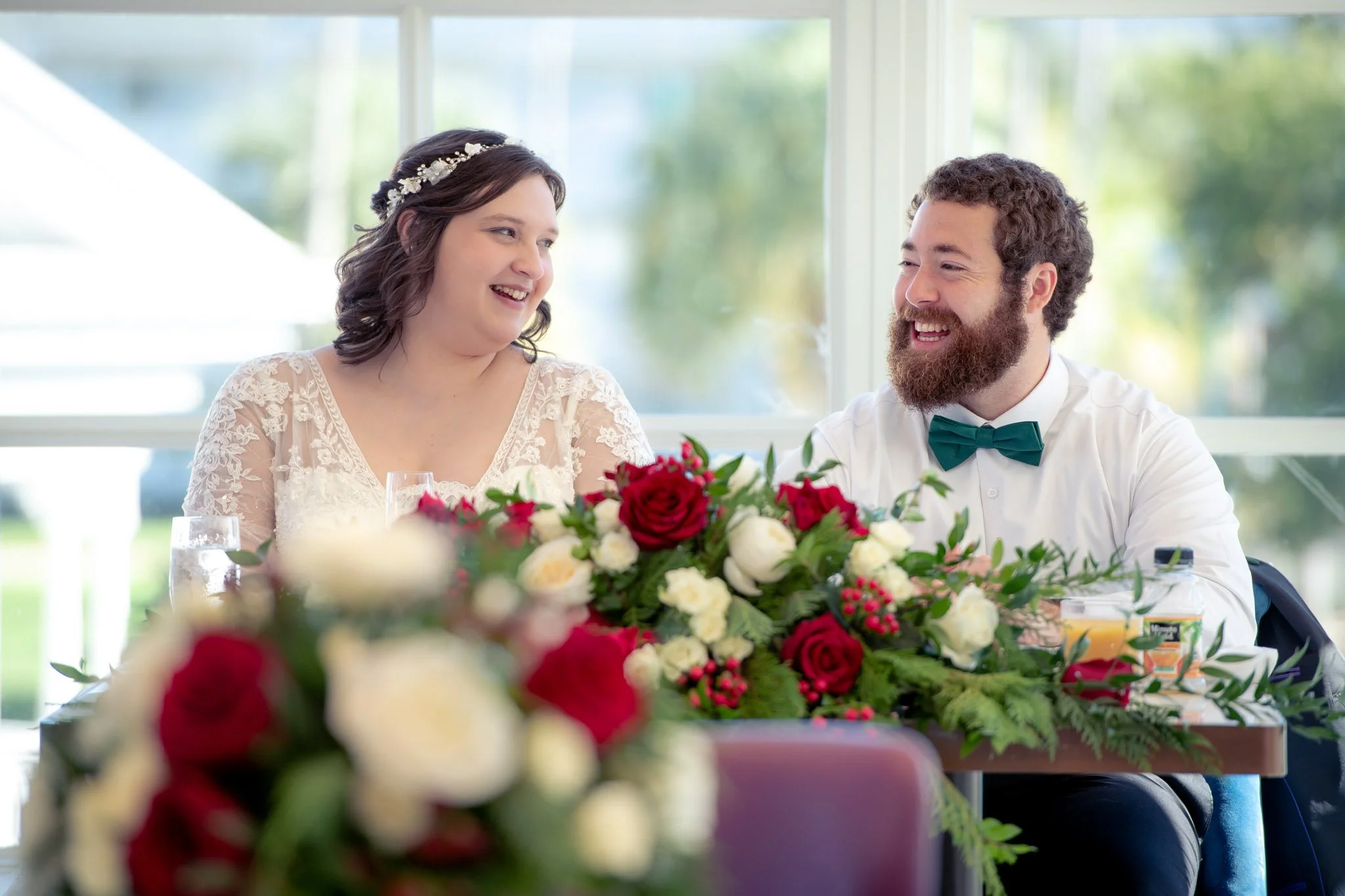 A bride and groom smiling during their wedding reception, sitting at a table decorated with a large floral arrangement of red and white roses.