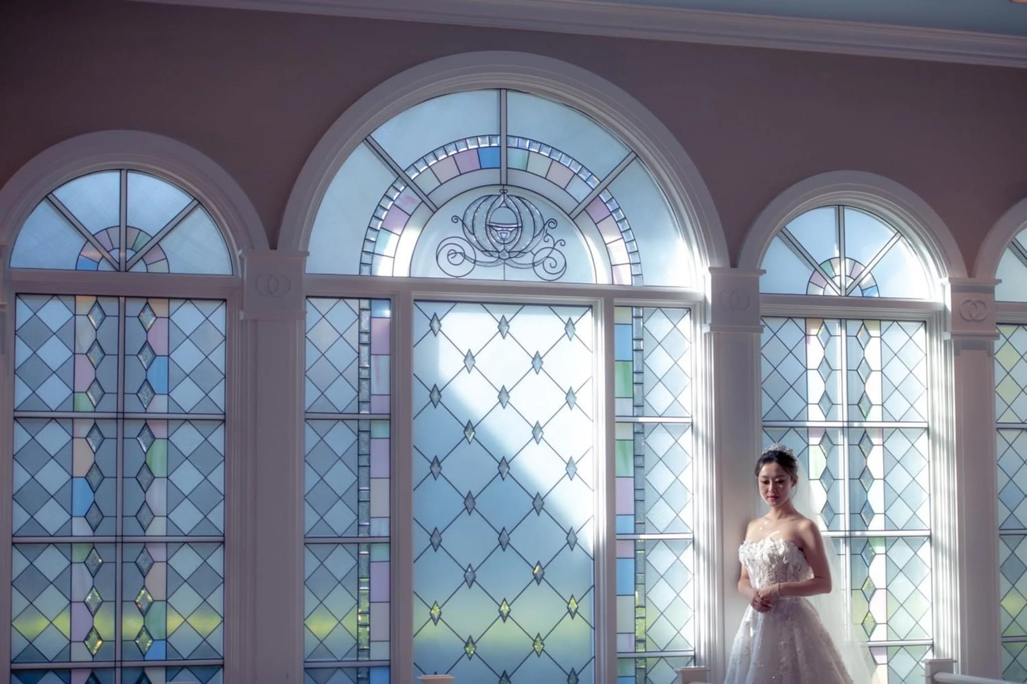 A woman in a wedding dress standing inside a brightly lit room at Disney's Wedding Pavilion with large stained glass windows.