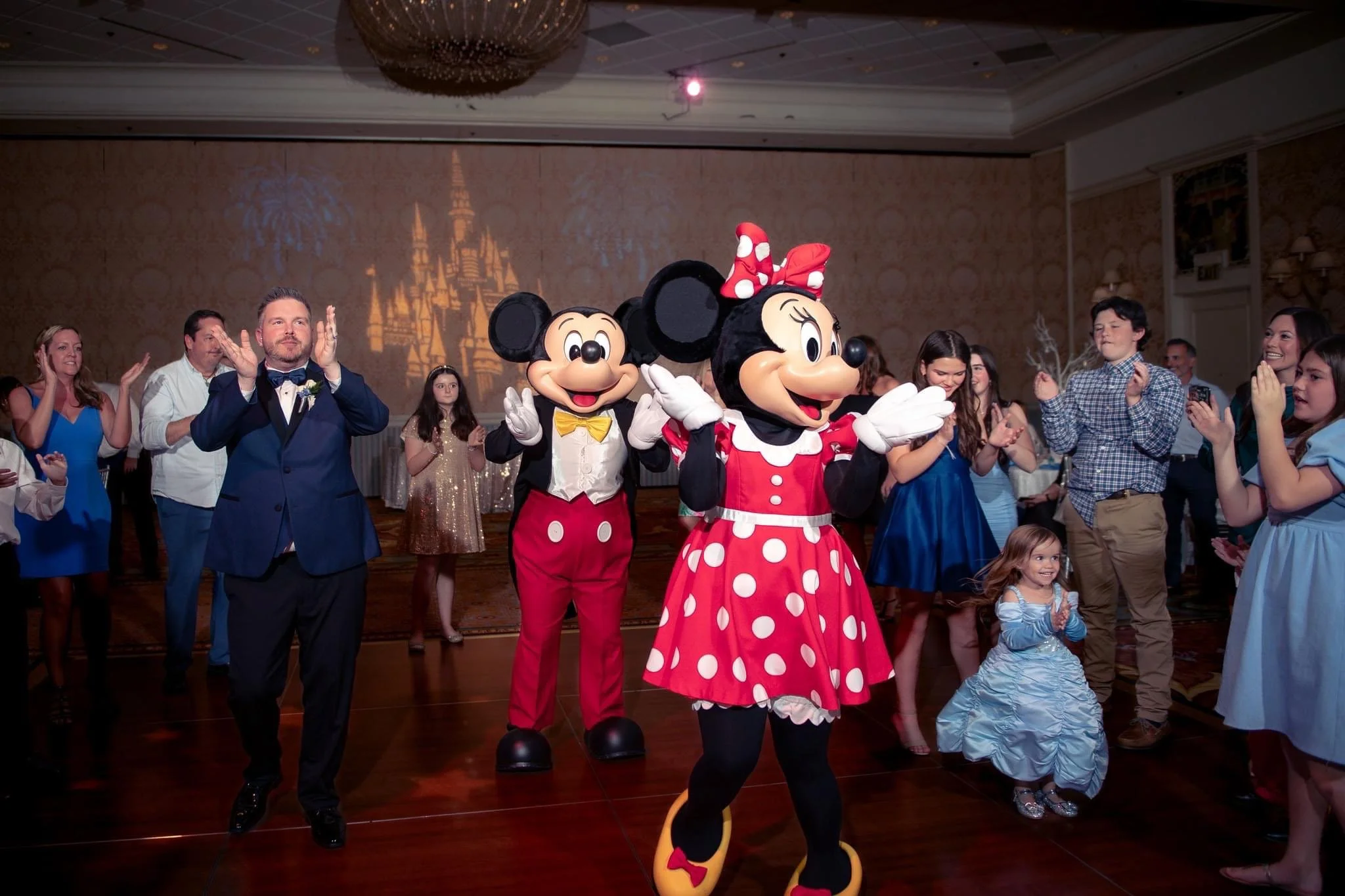 Guests at a party dancing with Mickey Mouse and Minnie Mouse, who are dressed in classic costumes, in a hotel banquet room with a projected castle shadow on the wall.