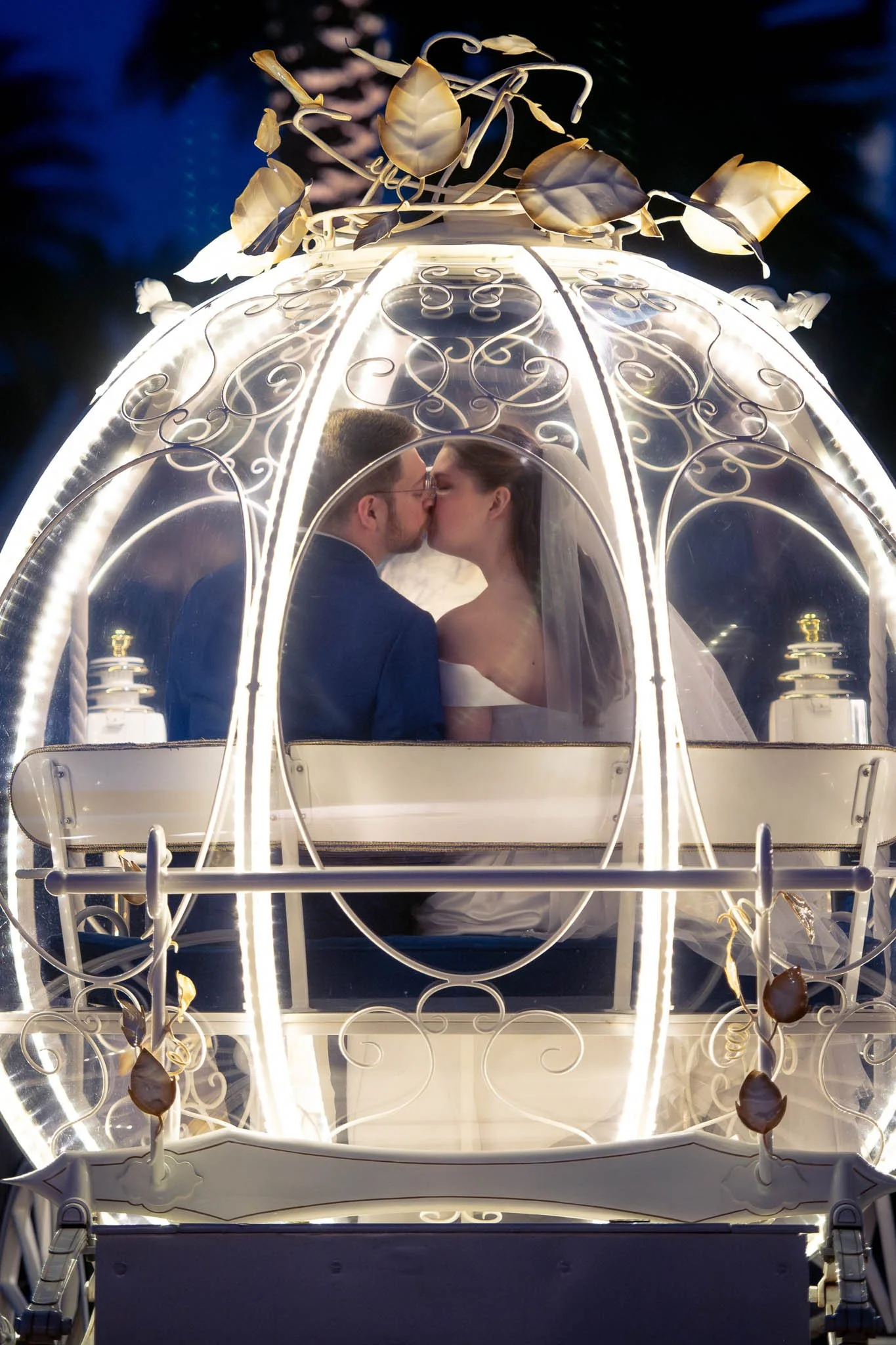 A bride and groom sharing a kiss inside a decorative, illuminated carriage at night.
