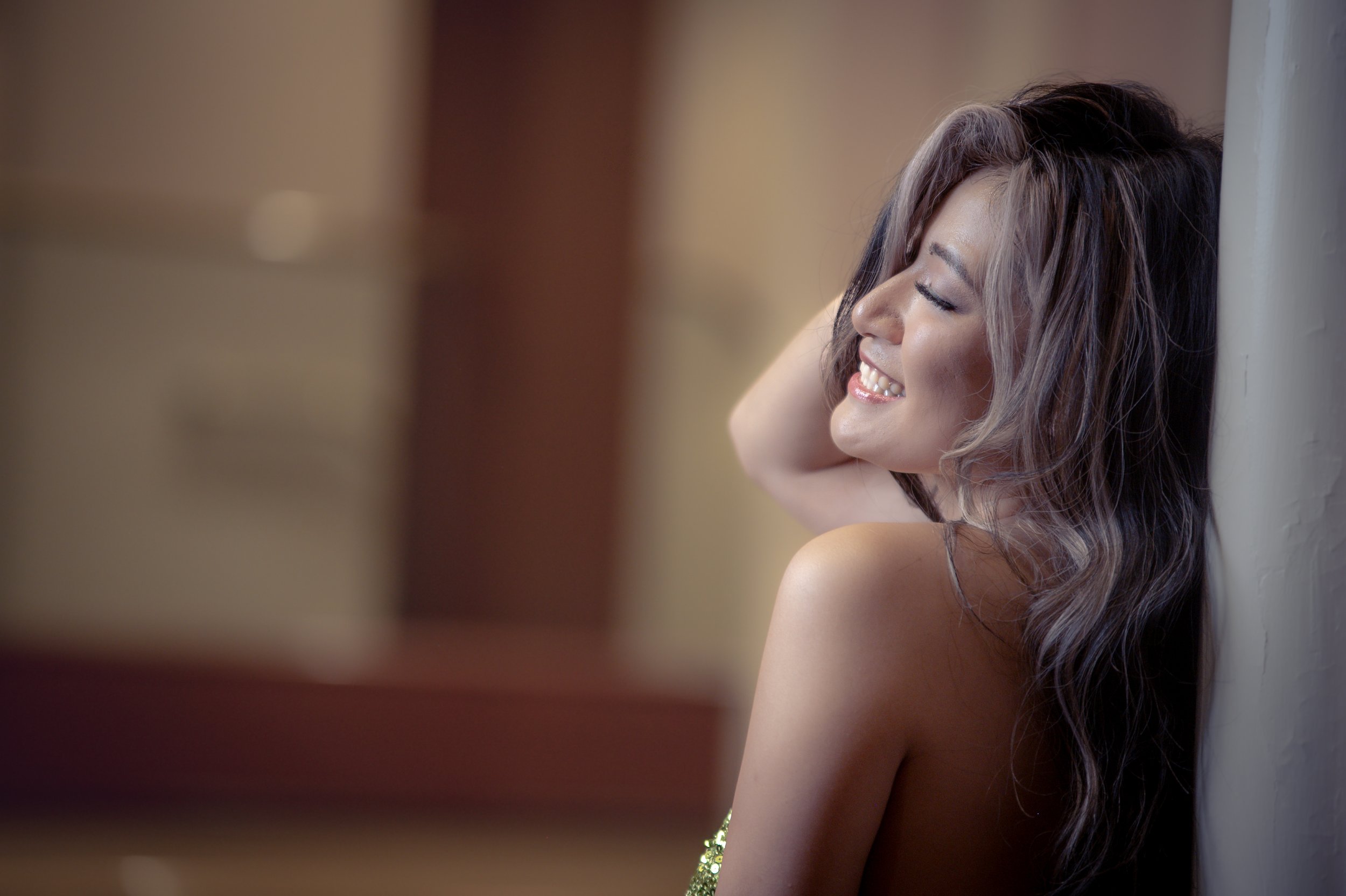 A woman with long wavy hair leaning back against a wall with her eyes closed, smiling, and touching her head with her hand.