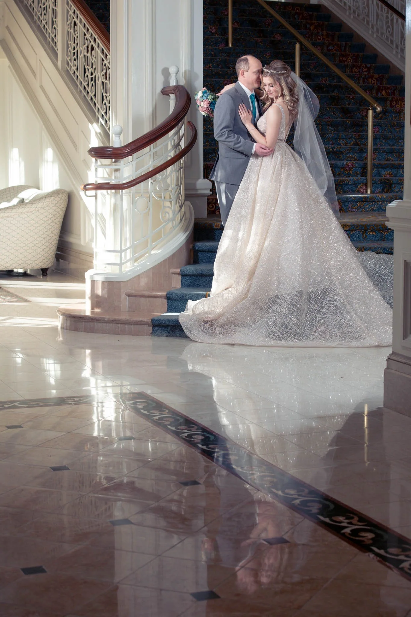 A bride and groom standing on a staircase, embracing and looking at each other. The bride wears a glittery wedding gown with a veil, and the groom wears a gray suit with a tie. The setting is indoors with polished marble floors, a curved staircase wi