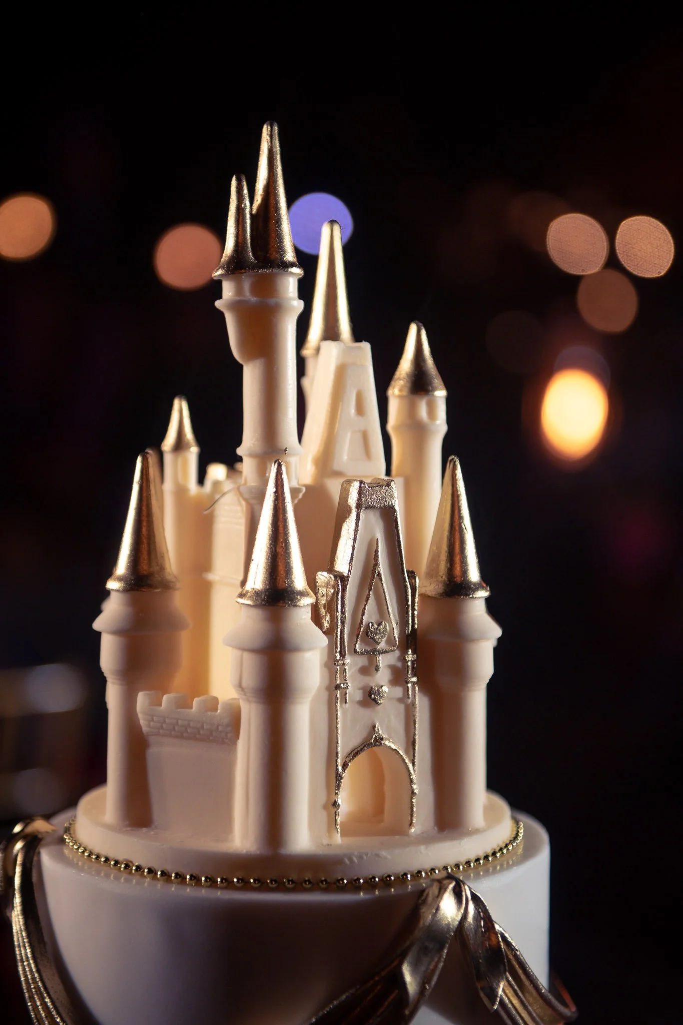 Decorative wedding cake shaped like a castle with white walls and gold accents on the towers, set against a dark background with bokeh lights.