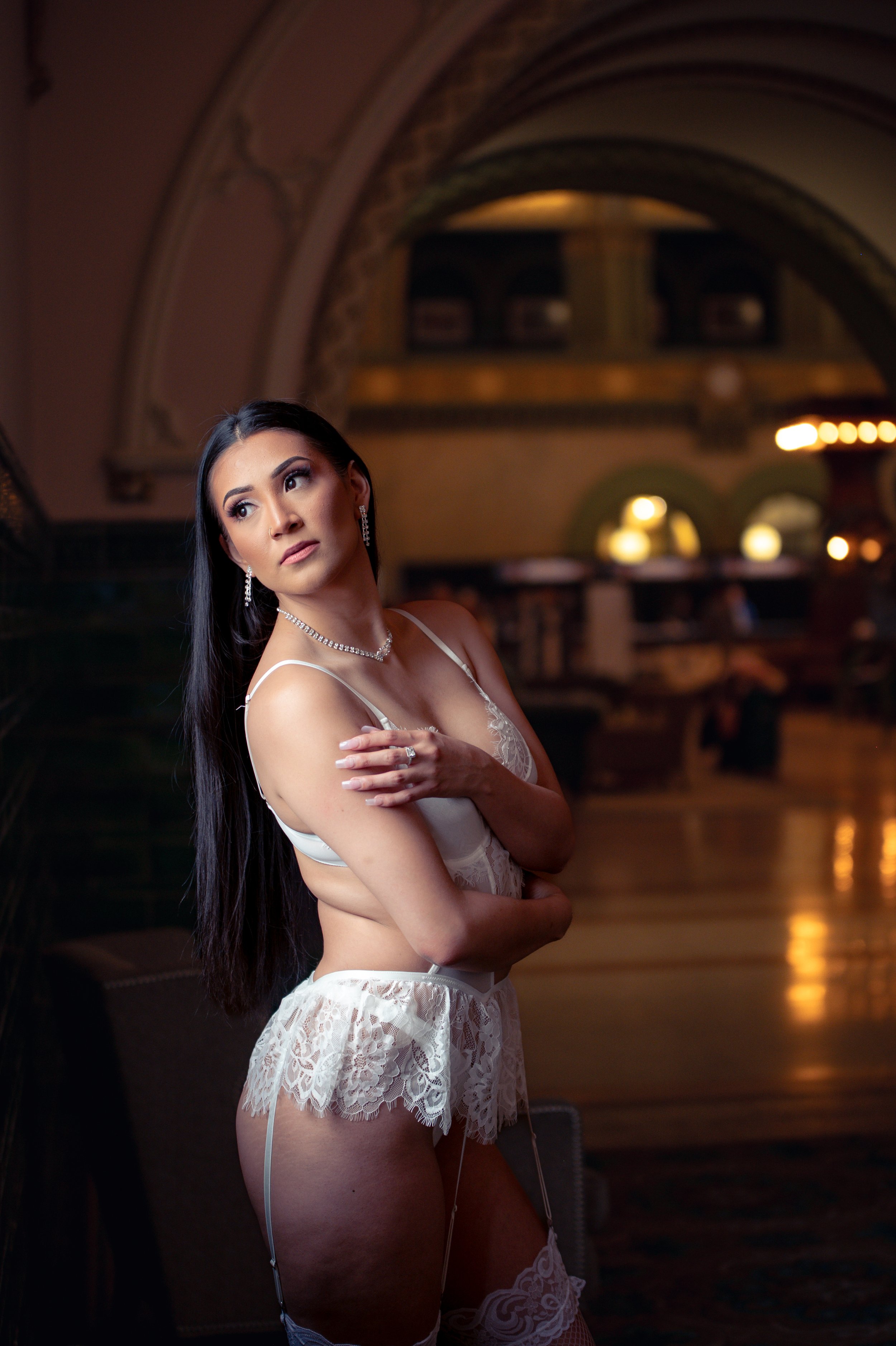 A woman with long black hair and jewelry, wearing white lingerie, posing in an elegant, dimly lit interior with arches and warm lighting.