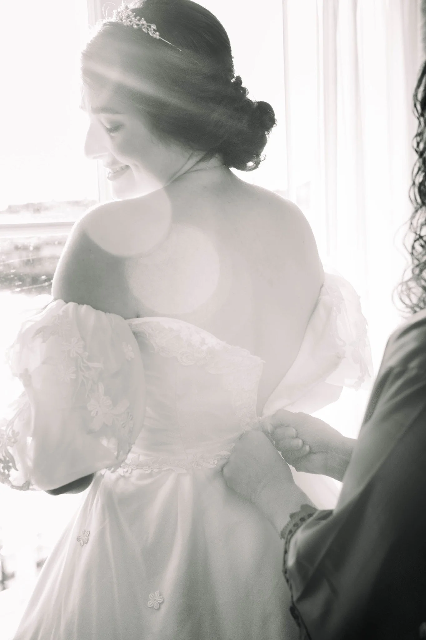 A woman in a wedding dress is being assisted with her dress by another person as she smiles with her eyes closed.