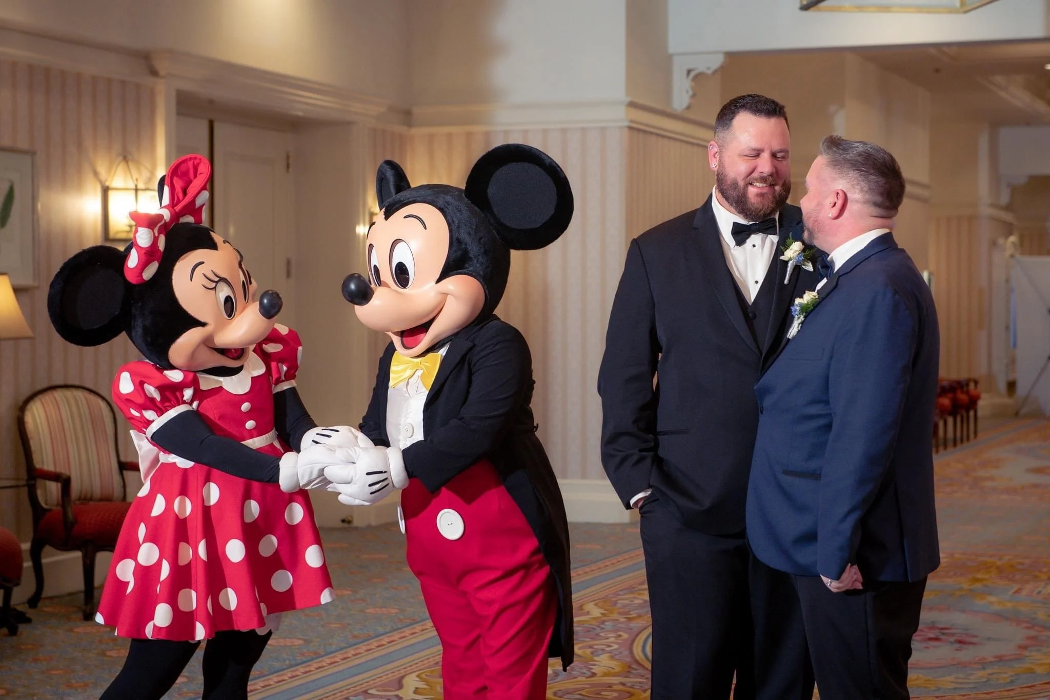 Two men in tuxedos, one with a beard, laughing and leaning close, are at a wedding reception. Two people in Mickey and Minnie Mouse costumes, in tuxedos and dresses, are holding hands near them, dancing or celebrating.