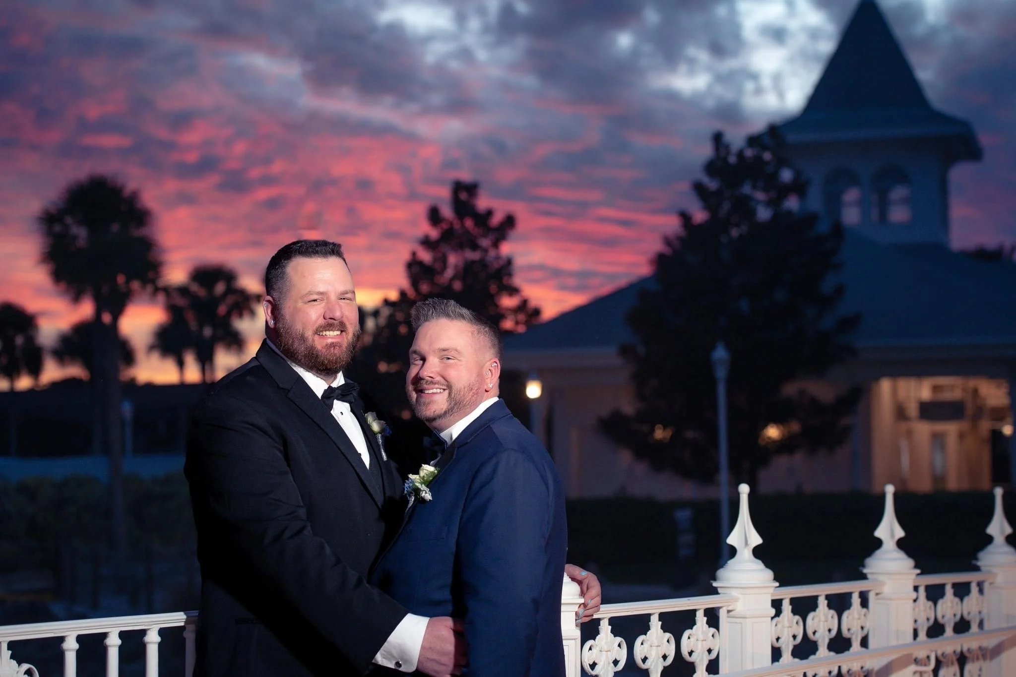 Two men in tuxedos at a wedding, smiling and embracing, with a colorful sunset in the background and a white decorative railing in the foreground.