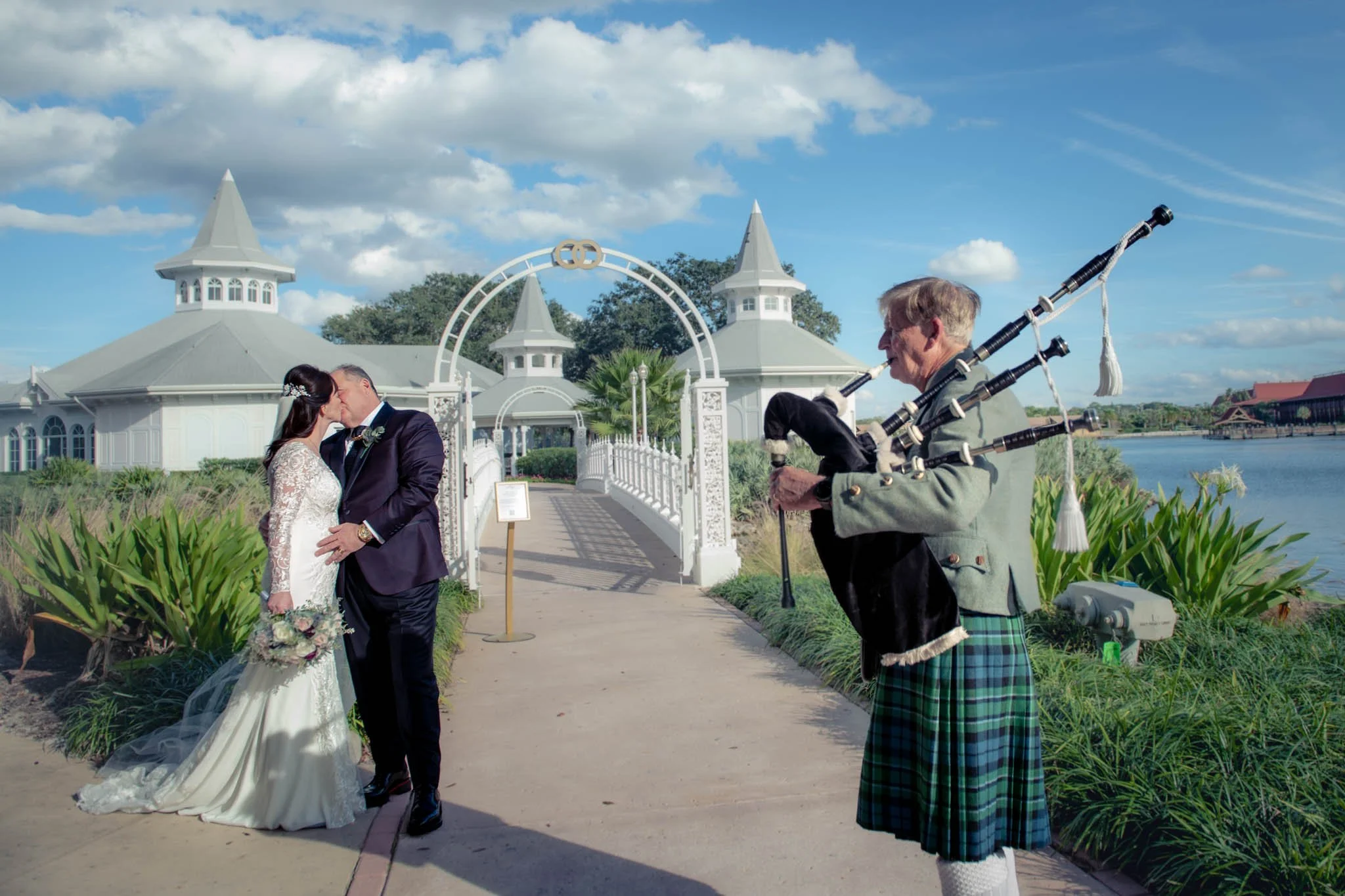 A couple getting married at Disney's Wedding Pavilion, sharing a kiss outdoors near a bridge and water, with a bagpiper playing nearby.
