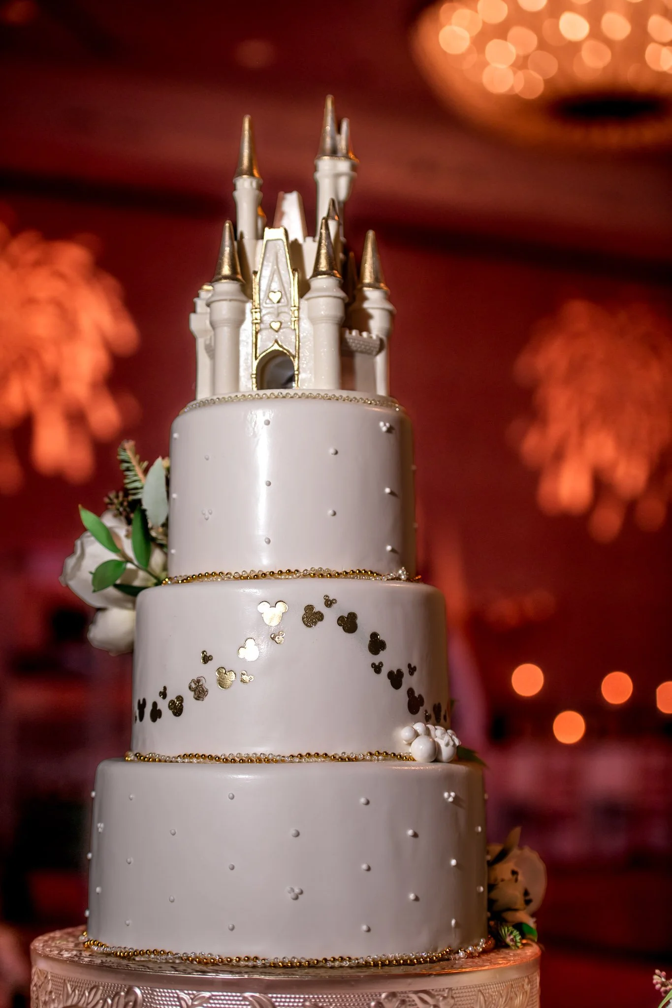 A large, elegant white wedding cake with four tiers decorated with small white and gold accents, featuring a castle-shaped topper. The cake is adorned with small flowers and greenery.