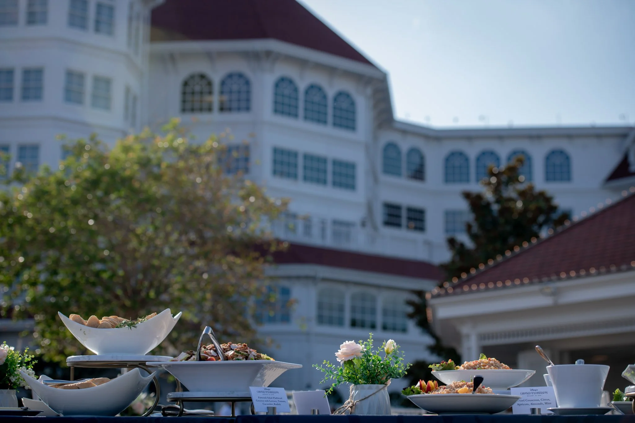 Outdoor buffet with bowls of salads and dishes, small potted flowers, and Disney's Grand Floridian with numerous windows in the background on a sunny day.