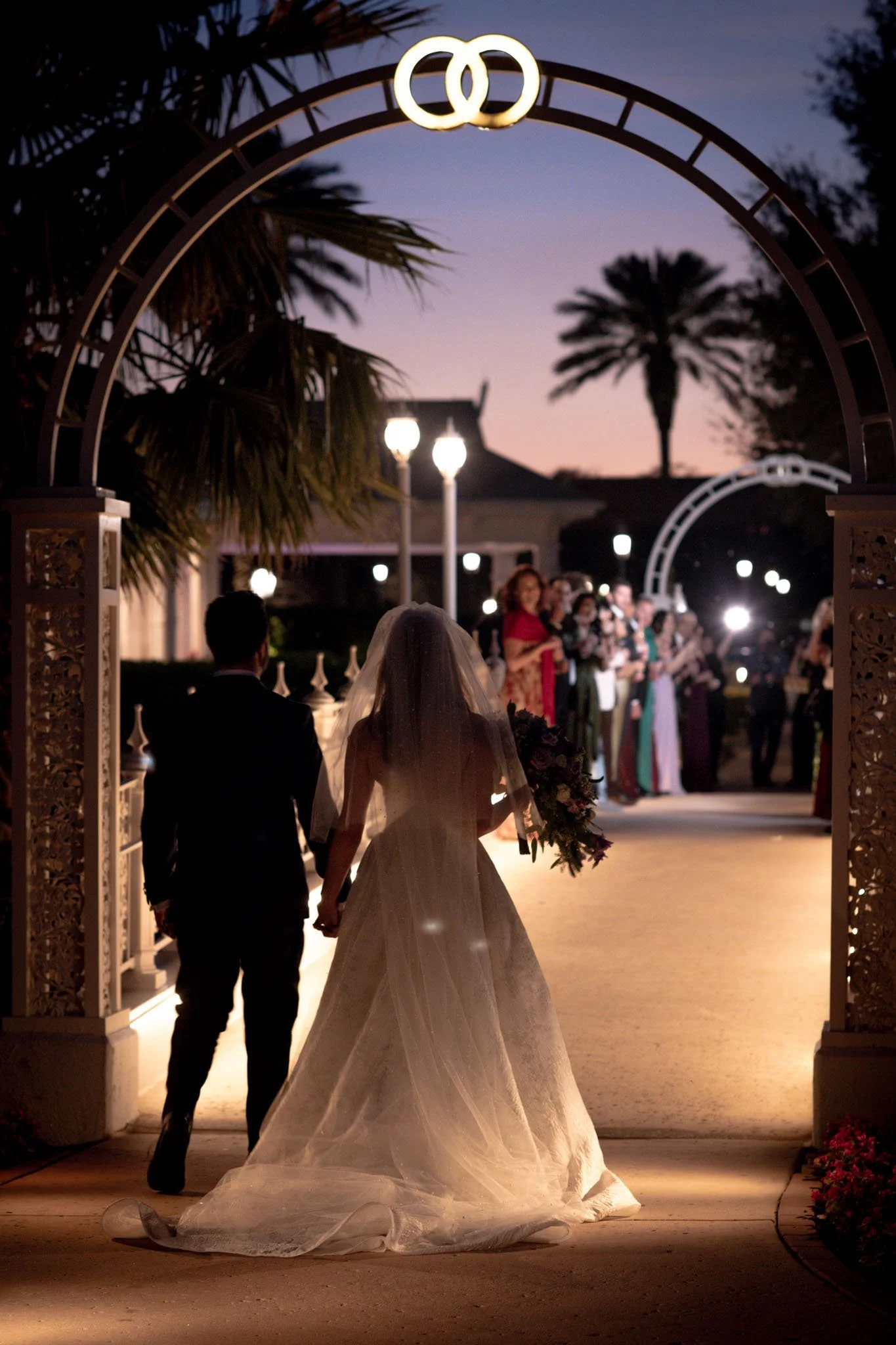 A bride and groom holding hands walking under an ornate arch at dusk, with guests lined up in the background and palm trees silhouetted against the evening sky.