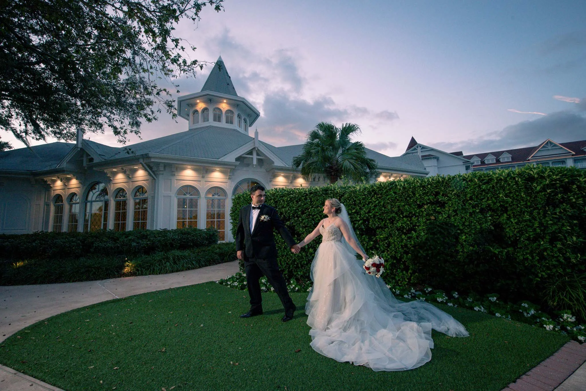 A newlywed couple holding hands outdoors at their wedding at Disney's Wedding Pavilion during sunset, with a large white building and hedge in the background.