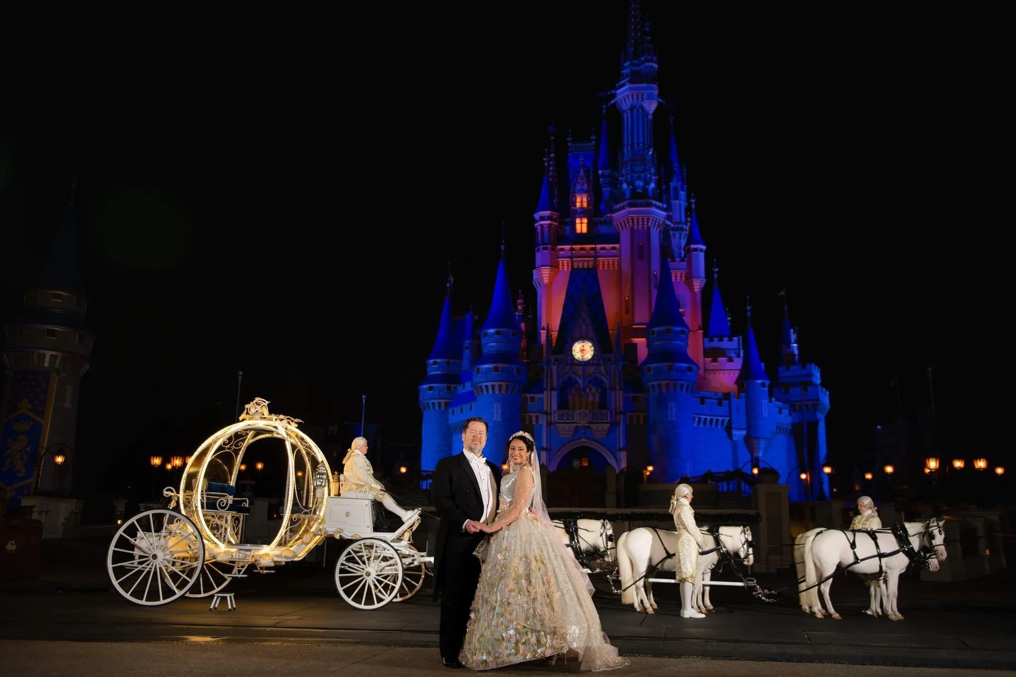 A couple in formal attire standing in front of Cinderella's castle at night, with a fairy tale carriage and horses in the background.