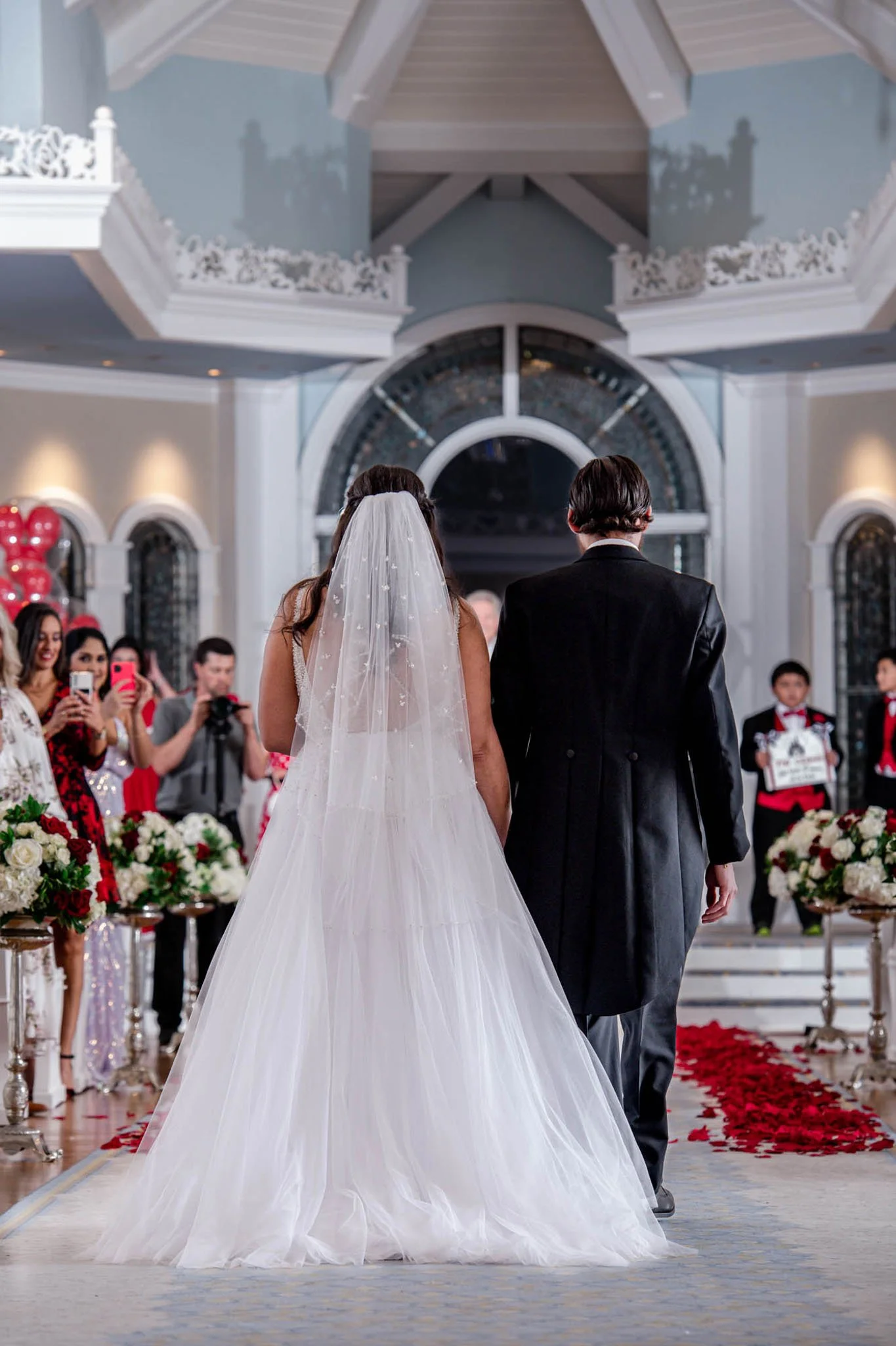 A bride in a white wedding gown and veil walking down the aisle with her groom in a black suit at a wedding ceremony holding hands, surrounded by guests and floral decorations.