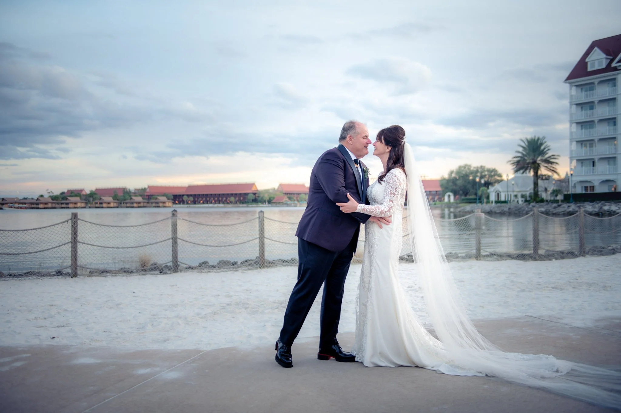 A bride and groom stand on a beach at The Grand Floridian, holding each other and smiling, with water and buildings in the background during sunset.