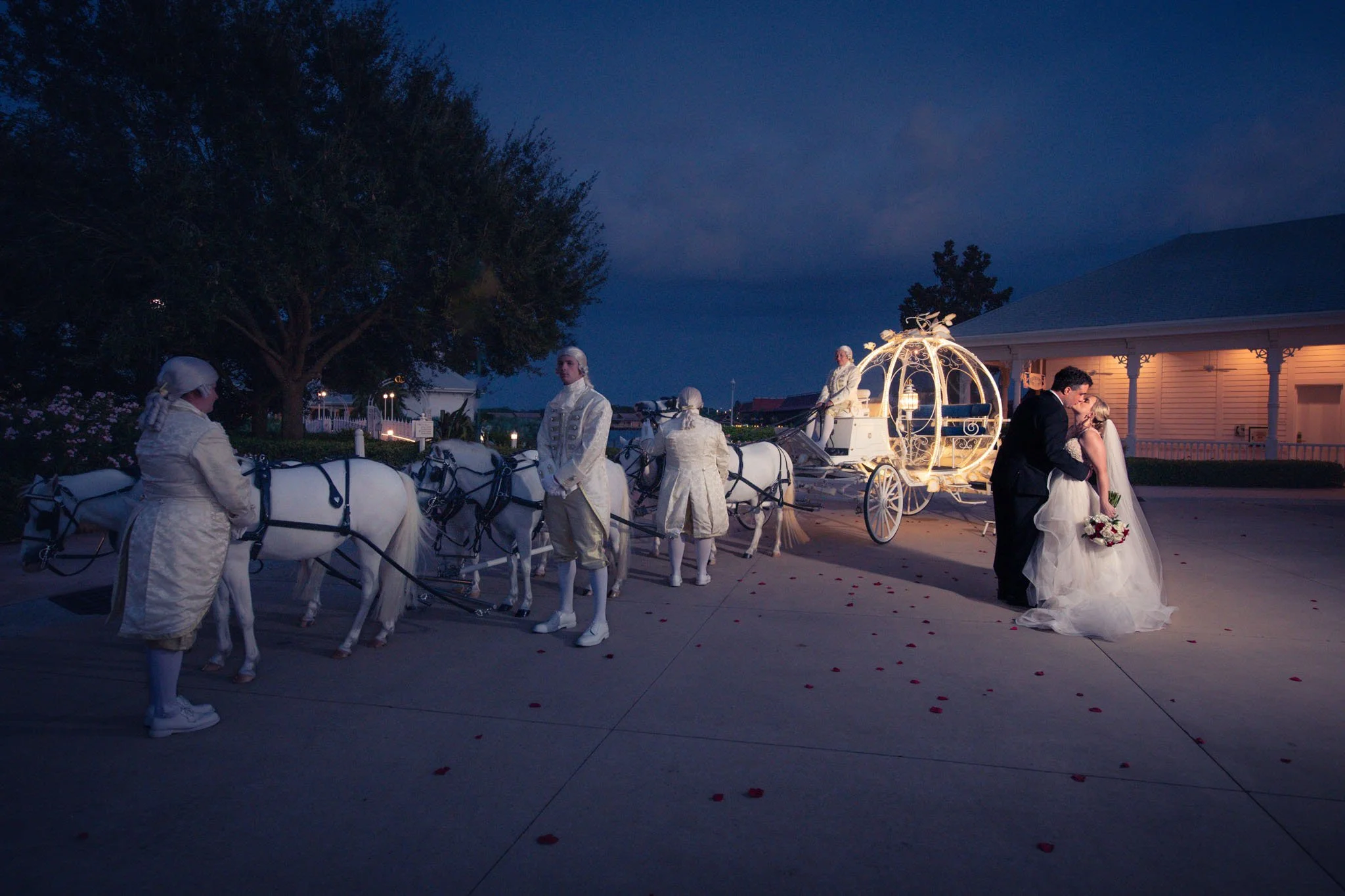 A wedding scene at night with a bride and groom sharing a kiss near a lit carriage and white horses, surrounded by people dressed in period costumes, outdoor setting with trees and a building in the background.