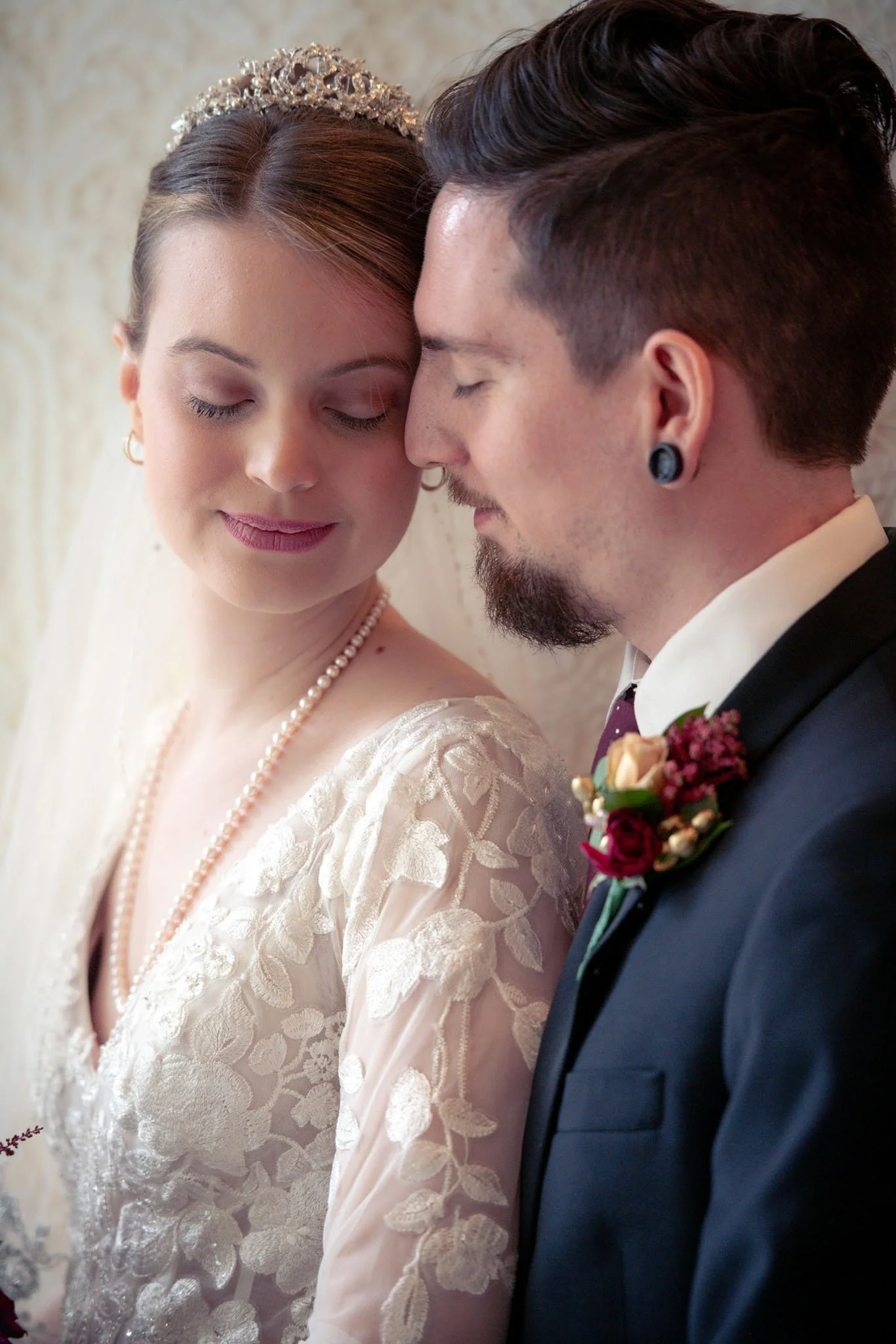 A bride and groom with foreheads touching, eyes closed, sharing an intimate moment at their wedding. The bride is wearing a lace dress, pearl necklace, and a tiara, while the groom is dressed in a tuxedo with a boutonnière.