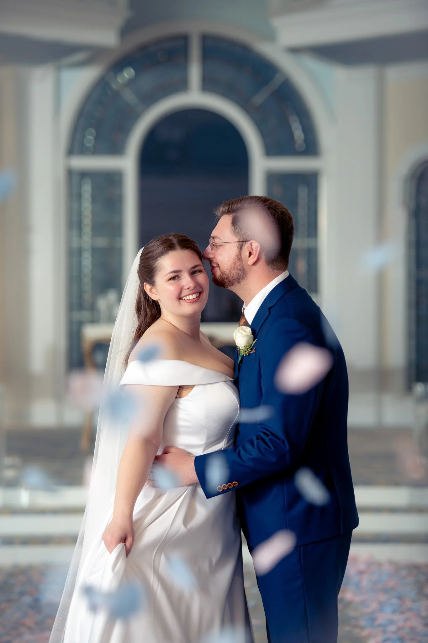 A newlywed couple in wedding attire, the bride in a white gown with a veil, and the groom in a navy suit, standing close together and smiling at each other, with blurred confetti falling around them.