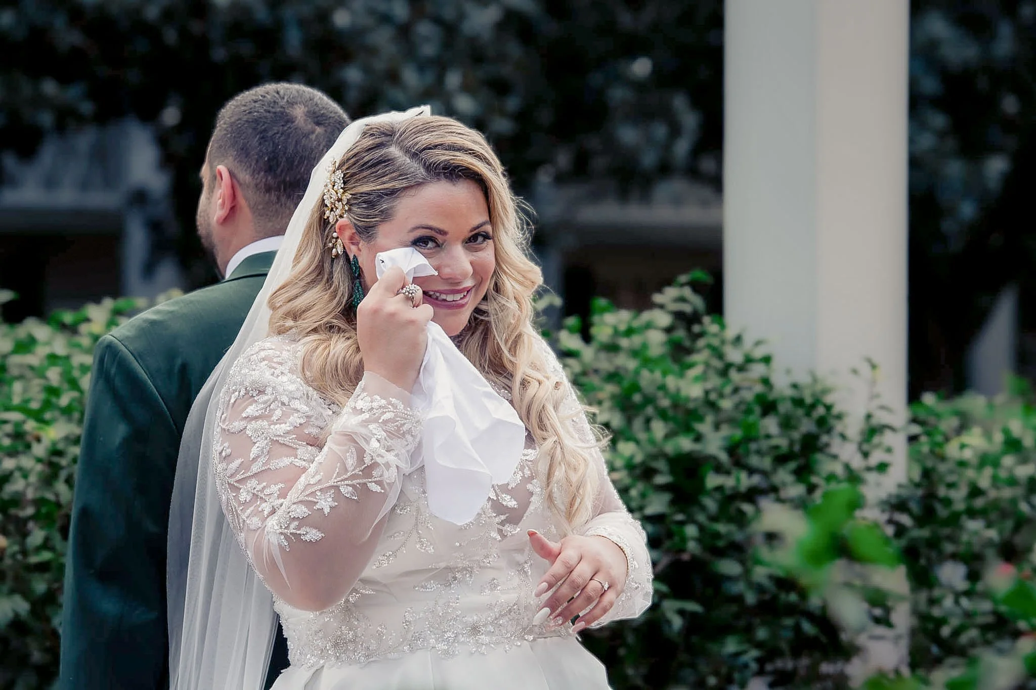 A bride with long curly hair wiping away tears with a tissue, smiling, wearing a lace wedding dress and veil, standing outdoors with a groom in a green suit behind her. Green bushes and trees in the background.