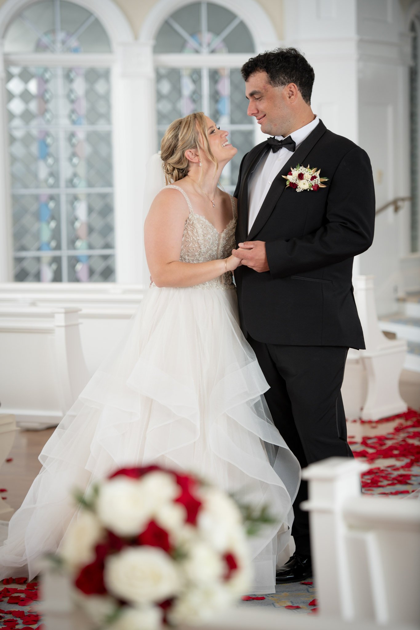 A bride and groom standing close together during their wedding ceremony in a bright, decorated church. The bride is in a white wedding gown with lace details and a veil, holding the groom's hands. The groom is dressed in a black tuxedo with a bow tie