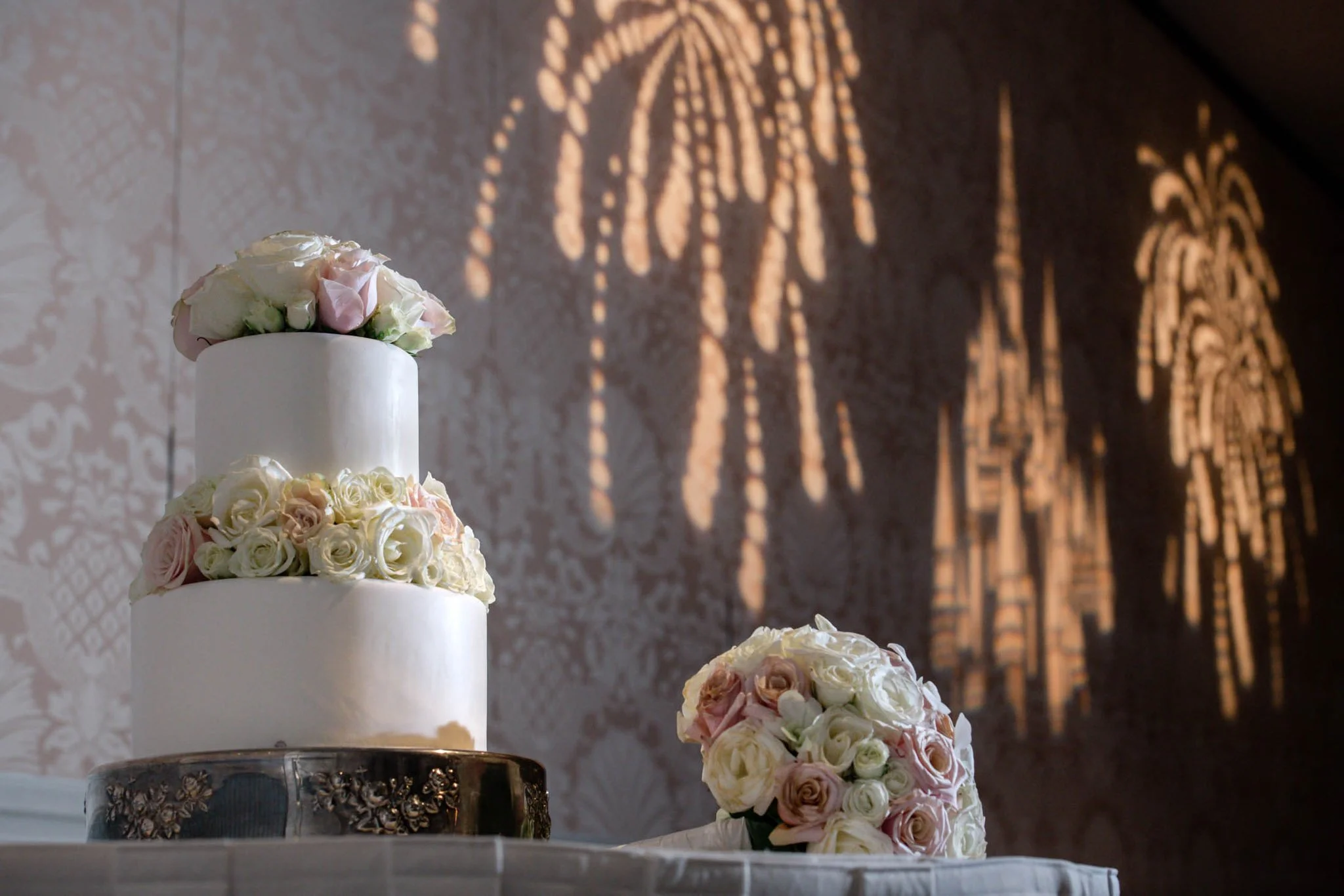 Three-tiered wedding cake decorated with white and light pink roses, placed on a table with a bouquet of roses nearby. Shadows of ornate decorations are cast on the wall in the background.