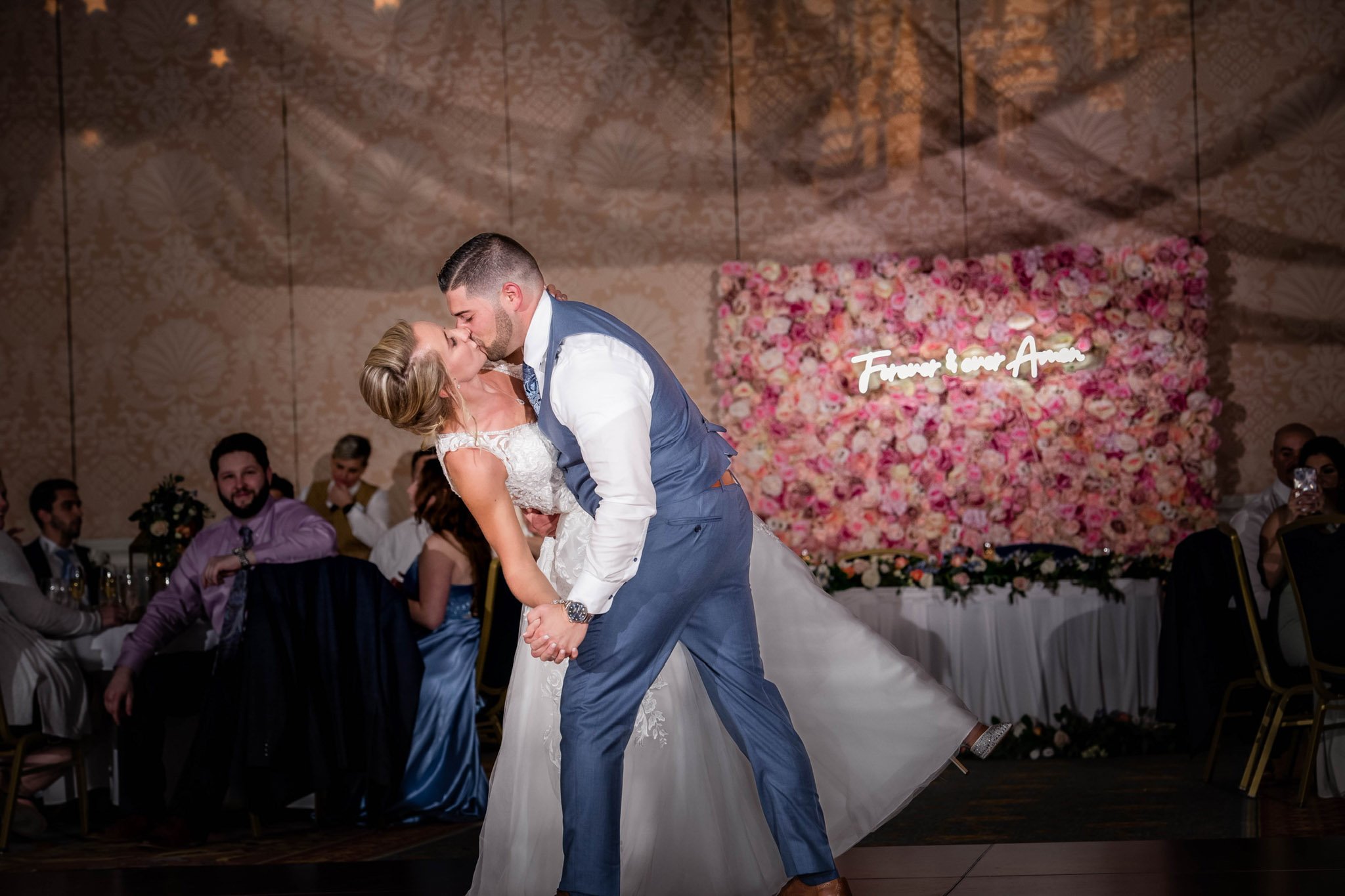 A newlywed couple sharing a kiss while dancing at their wedding reception, with guests seated at tables in the background and a floral backdrop with a neon sign reading 'Forever & Our Love'