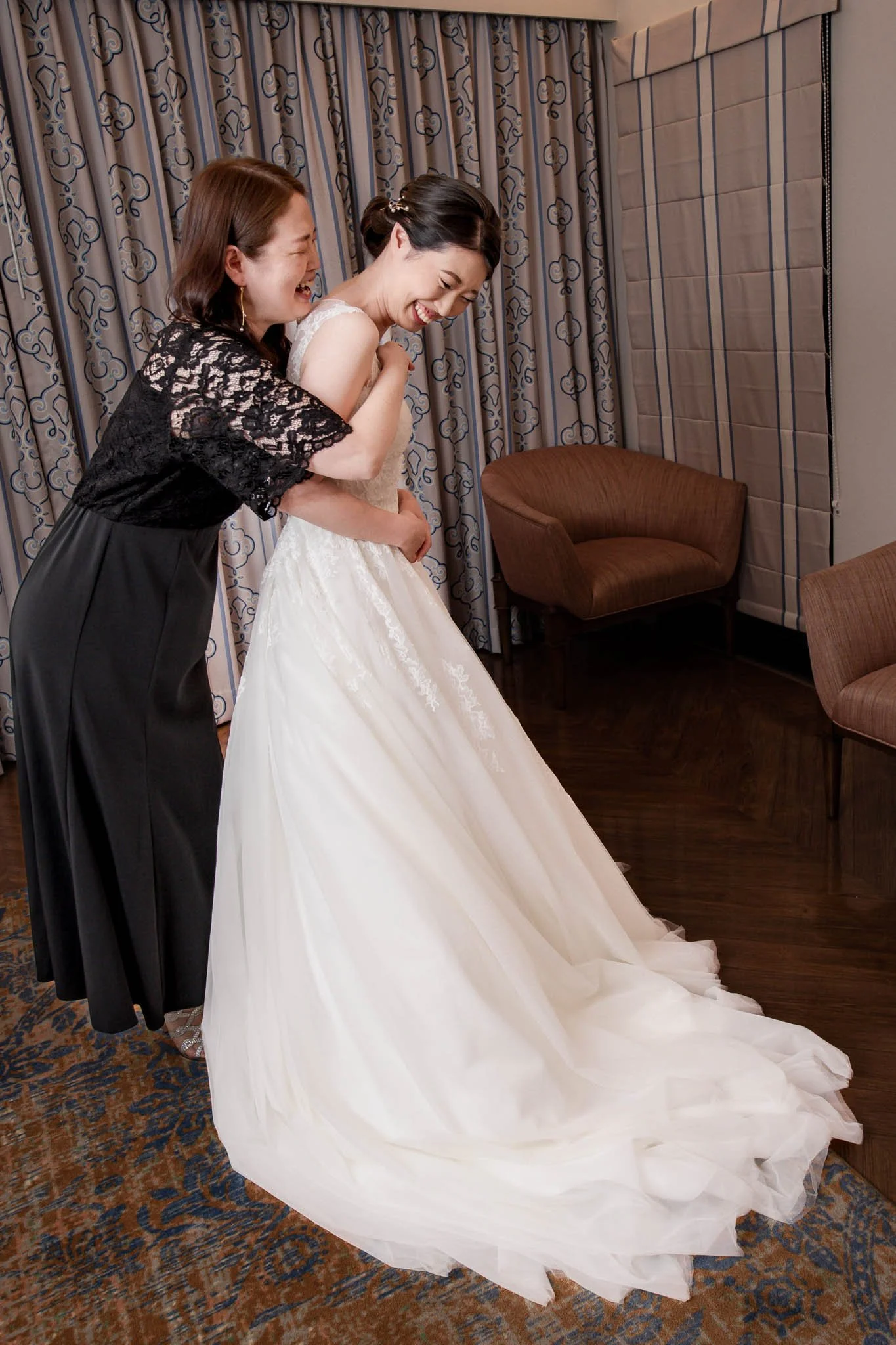 A bride in a white wedding dress and a woman in a black dress laughing and hugging each other in a room with patterned curtains and wooden furniture.