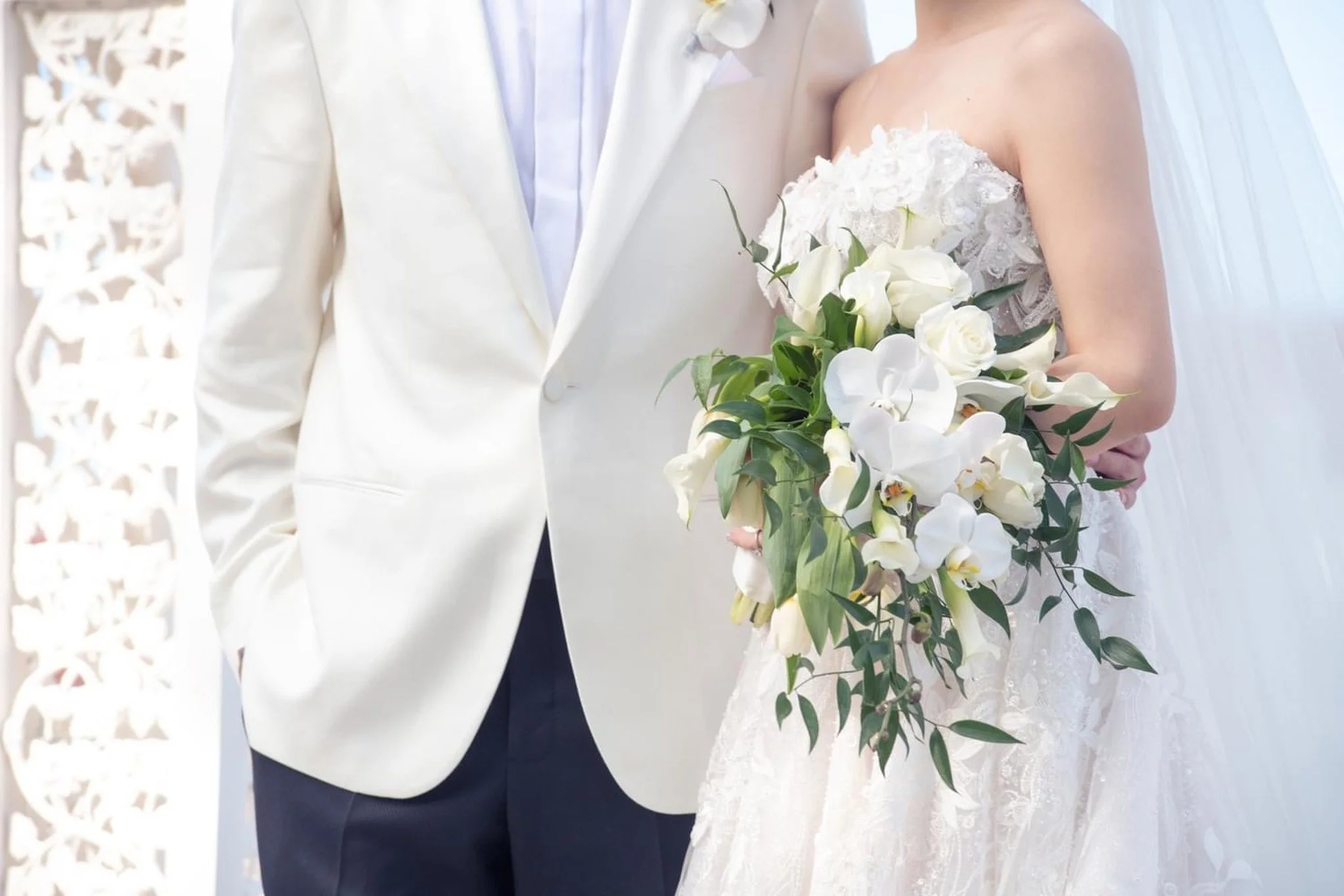 Close-up of a bride holding a bouquet of white flowers and a groom in a white suit at a wedding.