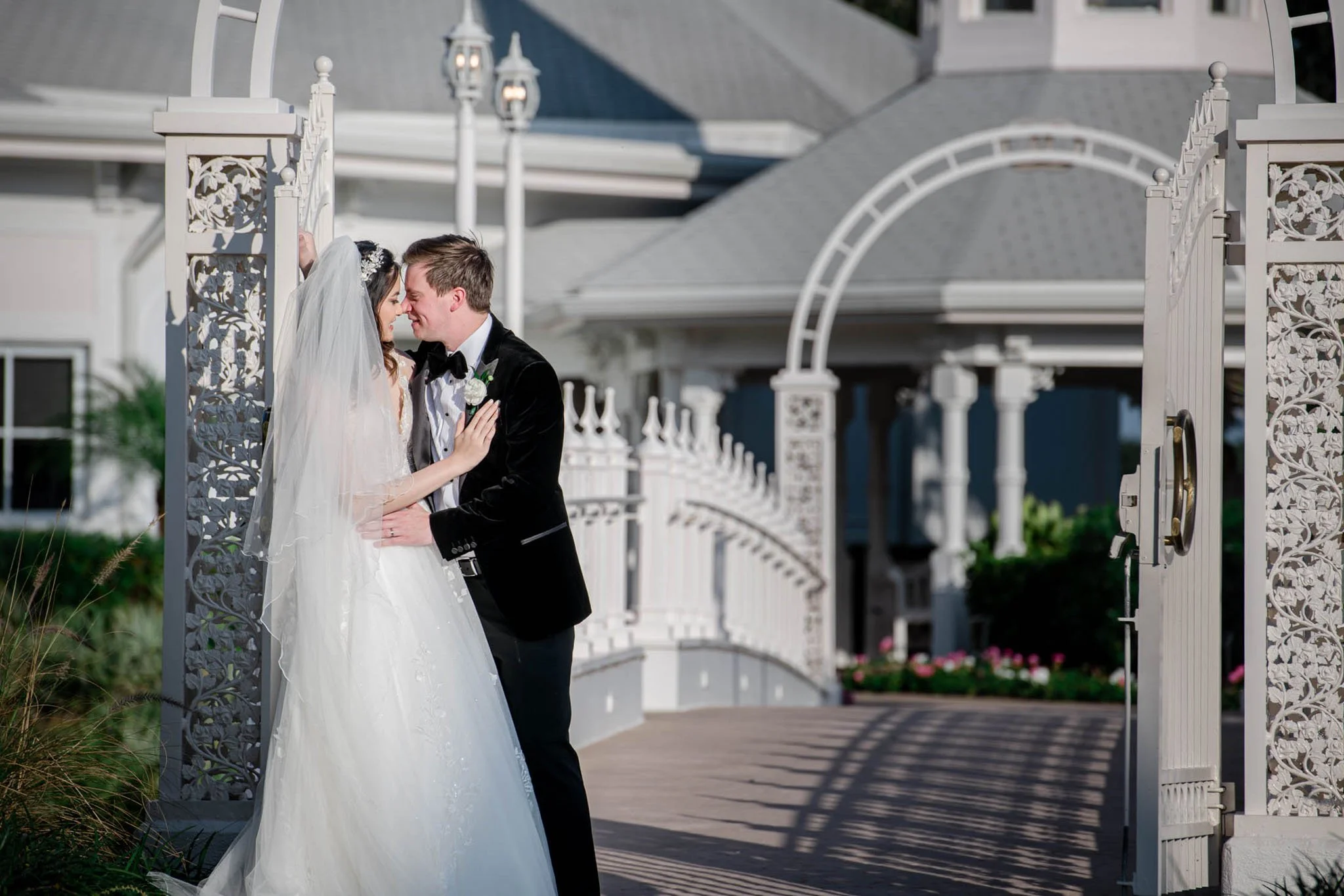 A bride and groom on their wedding day embracing under decorative white gate and archway, with Disney's Wedding Pavilion building in the background.
