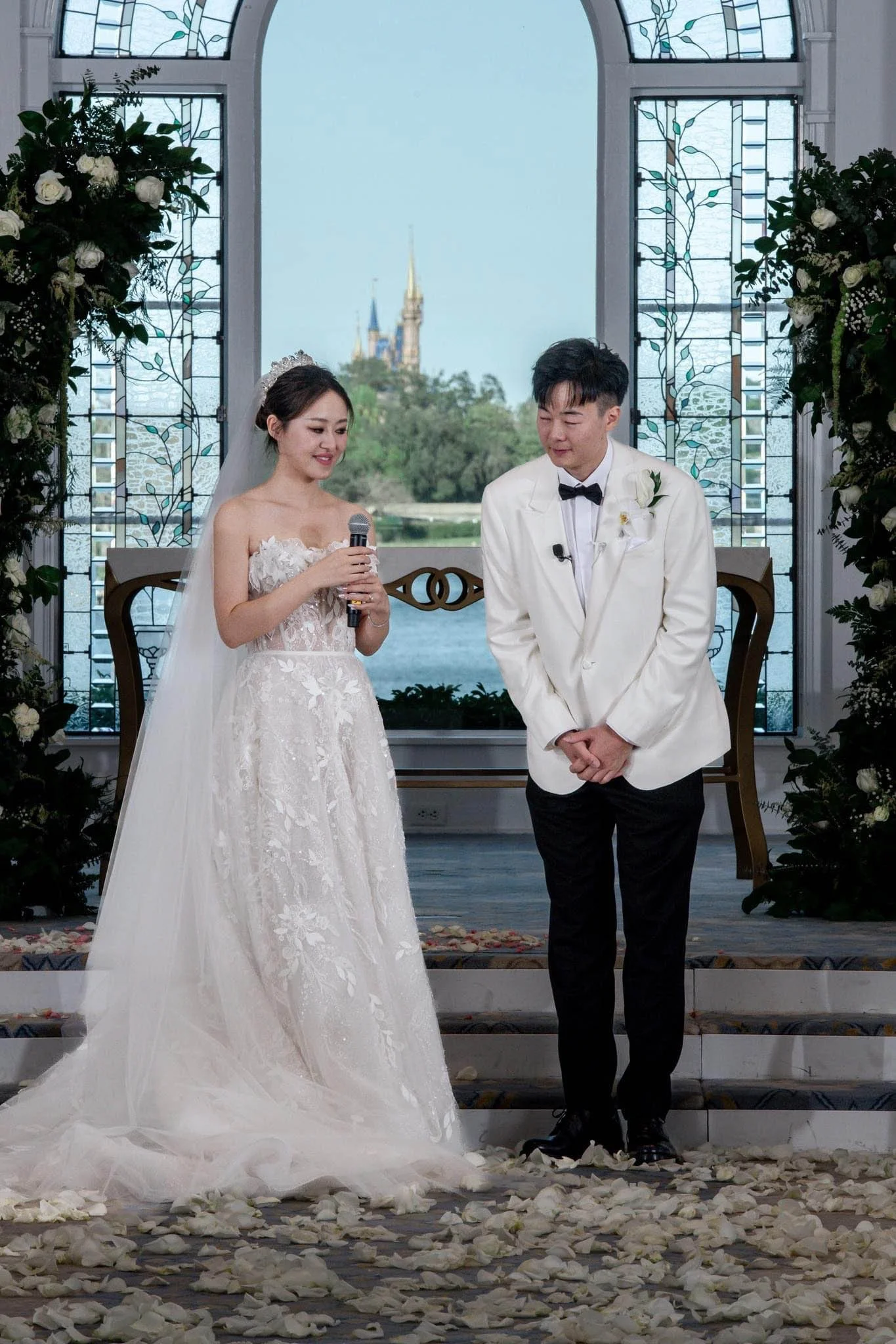 A bride and groom stand together in a wedding ceremony, with the bride holding a microphone. The bride wears a white wedding gown with lace details and a veil, while the groom wears a white tuxedo jacket with black pants and a bow tie. They are on a 