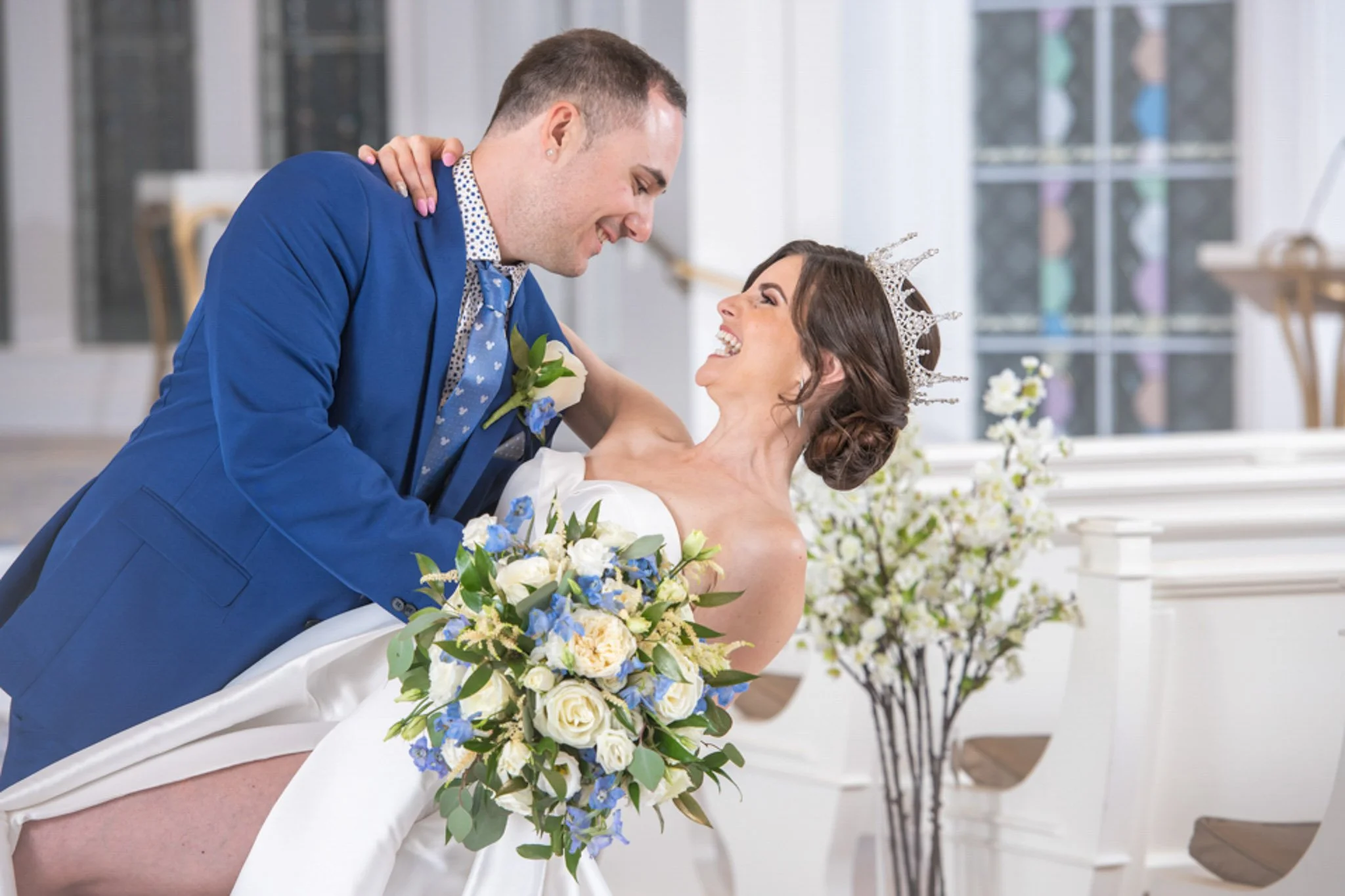 A couple dressed in wedding attire dancing and smiling indoors, with the woman holding a bouquet of white and blue flowers, and the man wearing a blue suit with a boutonniere.