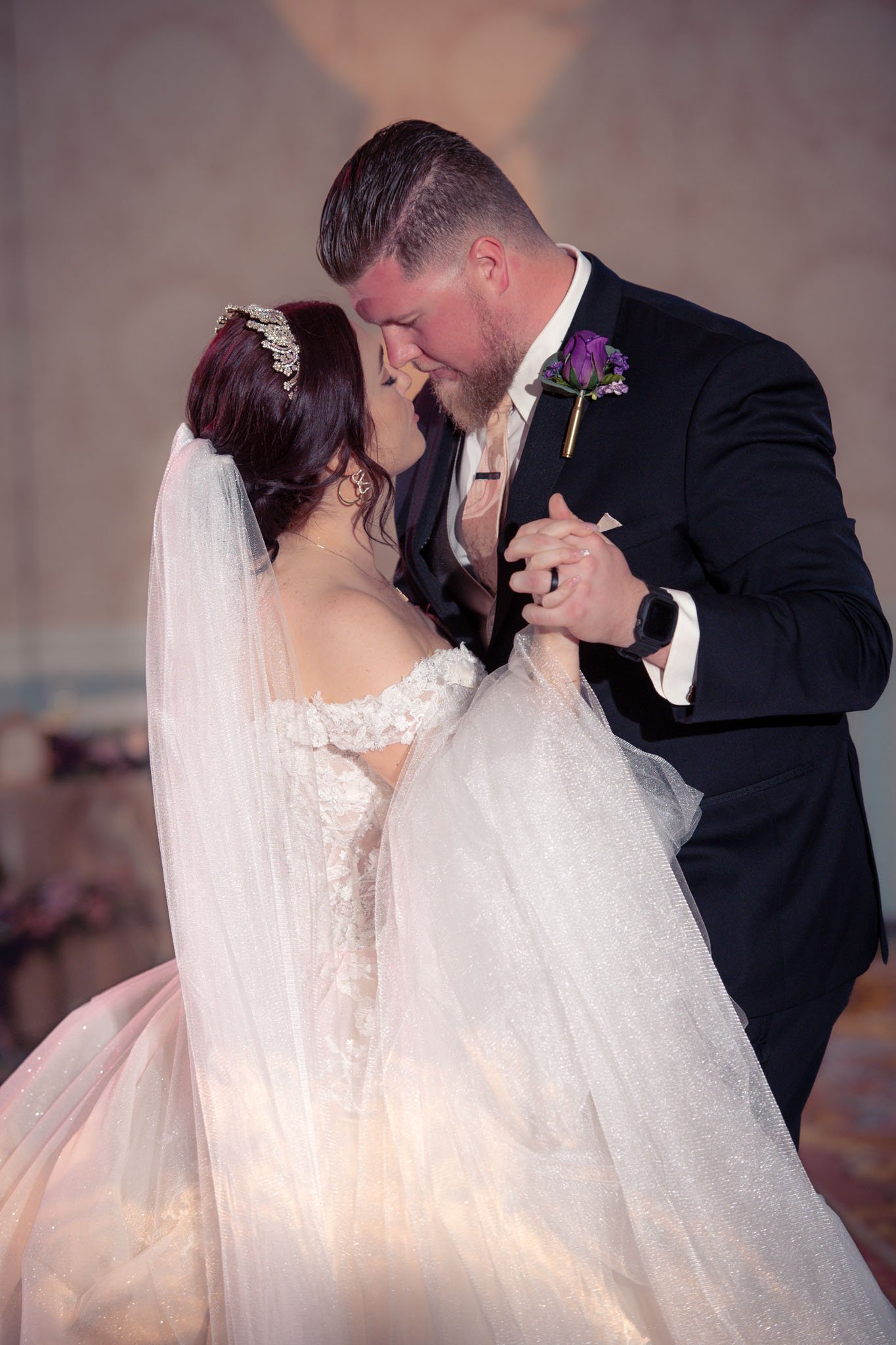 A bride and groom share a dance, with the bride wearing an off-shoulder wedding gown and veil, and the groom dressed in a dark suit with a purple boutonniere.