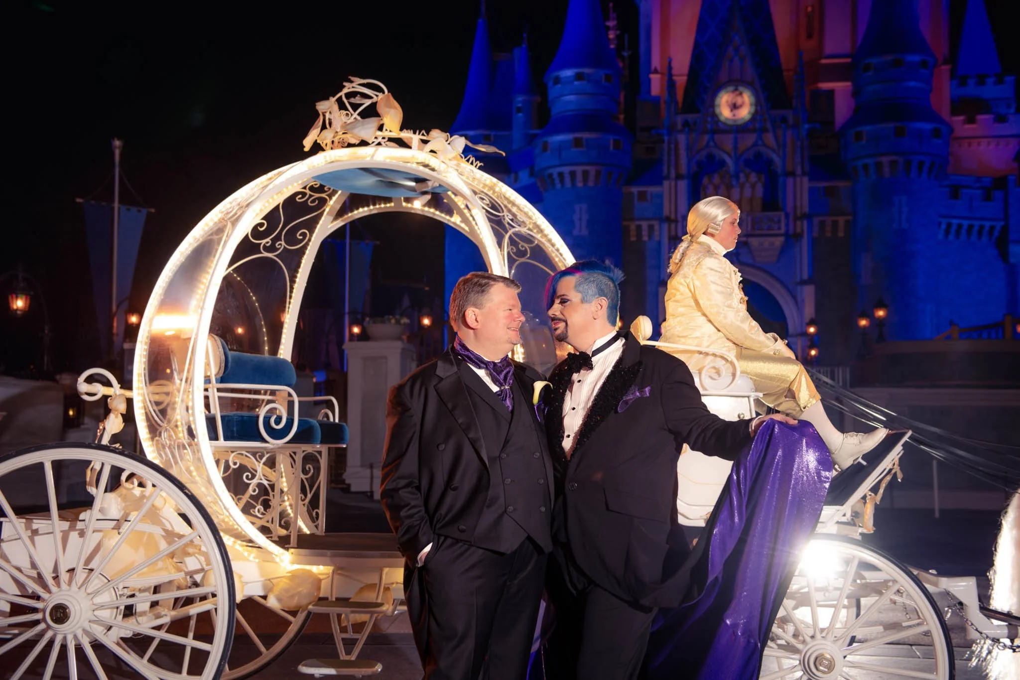 Two men in tuxedos standing close together in front of a Disney castle at night, with a fairy tale carriage at their Magic Kingdom Wedding.