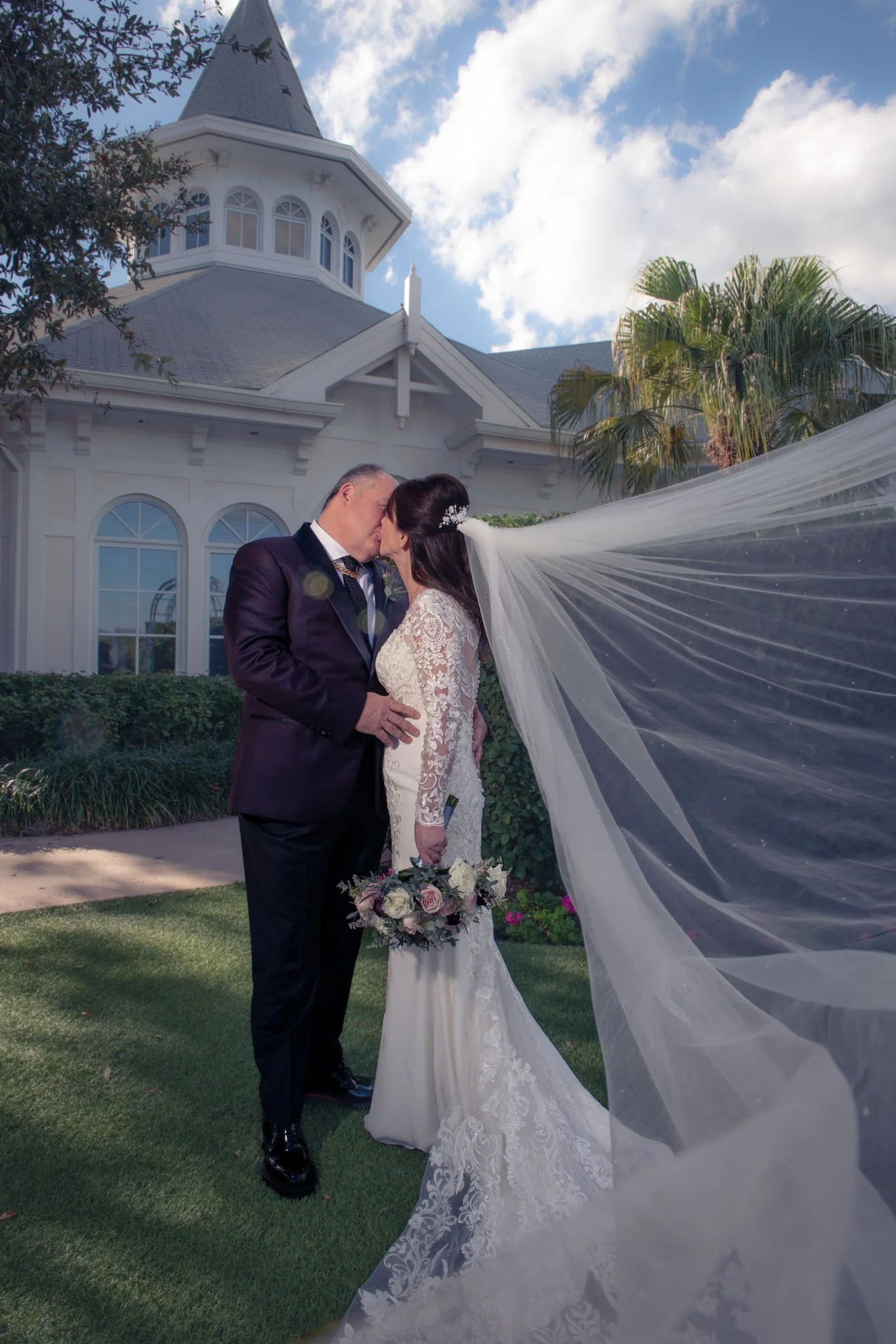 Bride and groom sharing a kiss outdoors on a sunny day, with a large white house and palm trees in the background. The bride wears a lace wedding dress and veil, holding a bouquet of pink and white flowers. The groom wears a dark suit.