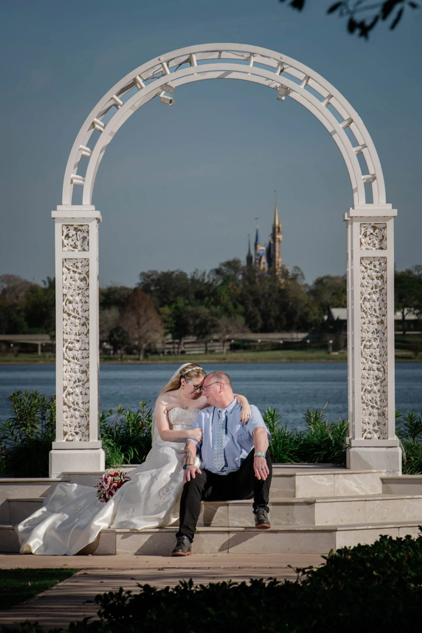 A wedding couple sitting on stairs under a decorative white arch near a body of water with Cinderella's castle in the background.