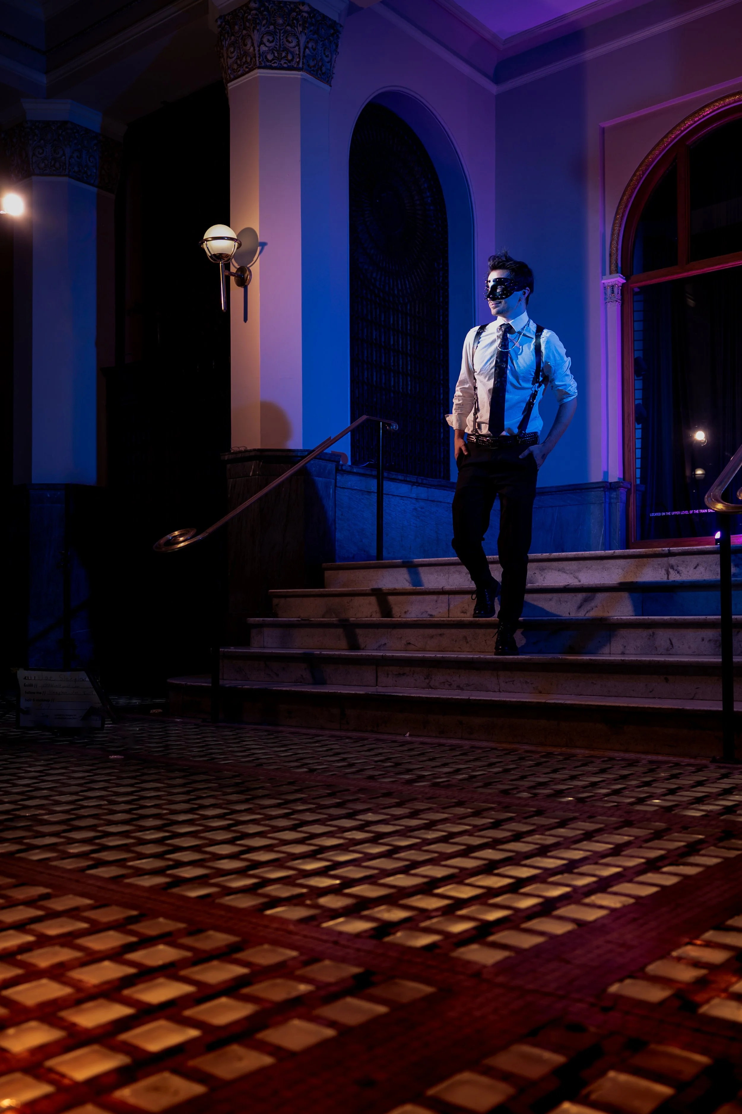 A person wearing a mask and formal attire walking down a staircase in a dimly lit, elegant building with purple and blue lighting.