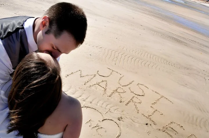A couple kissing on a beach with 'Just Married' written in the sand.
