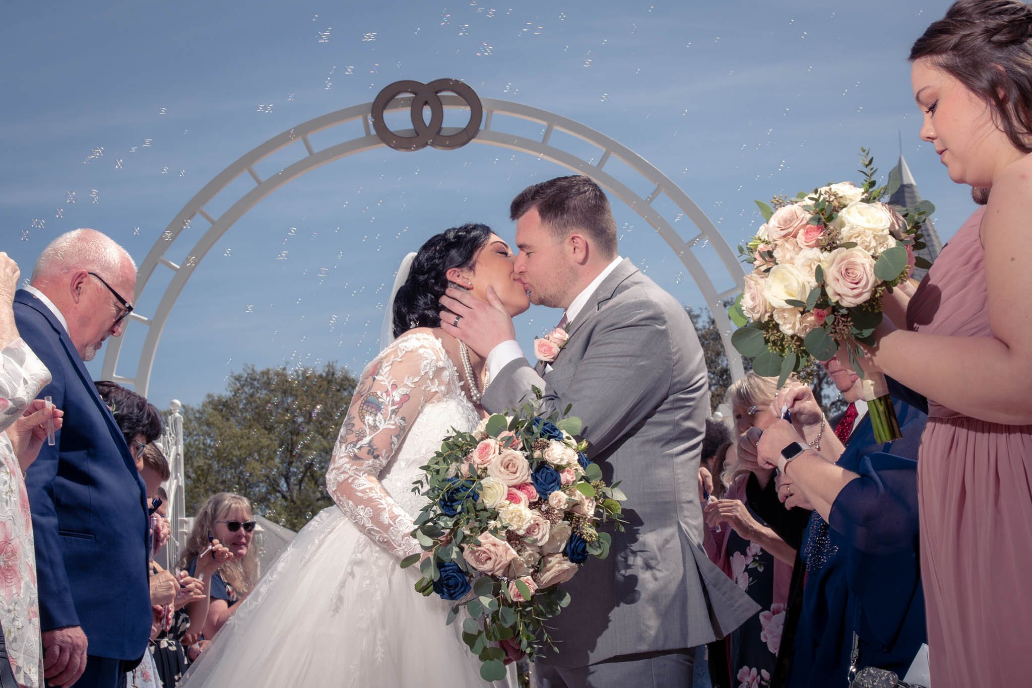 A couple getting married, kissing under a wedding arch with a small circle emblem, surrounded by wedding guests holding bouquets, on a sunny day.