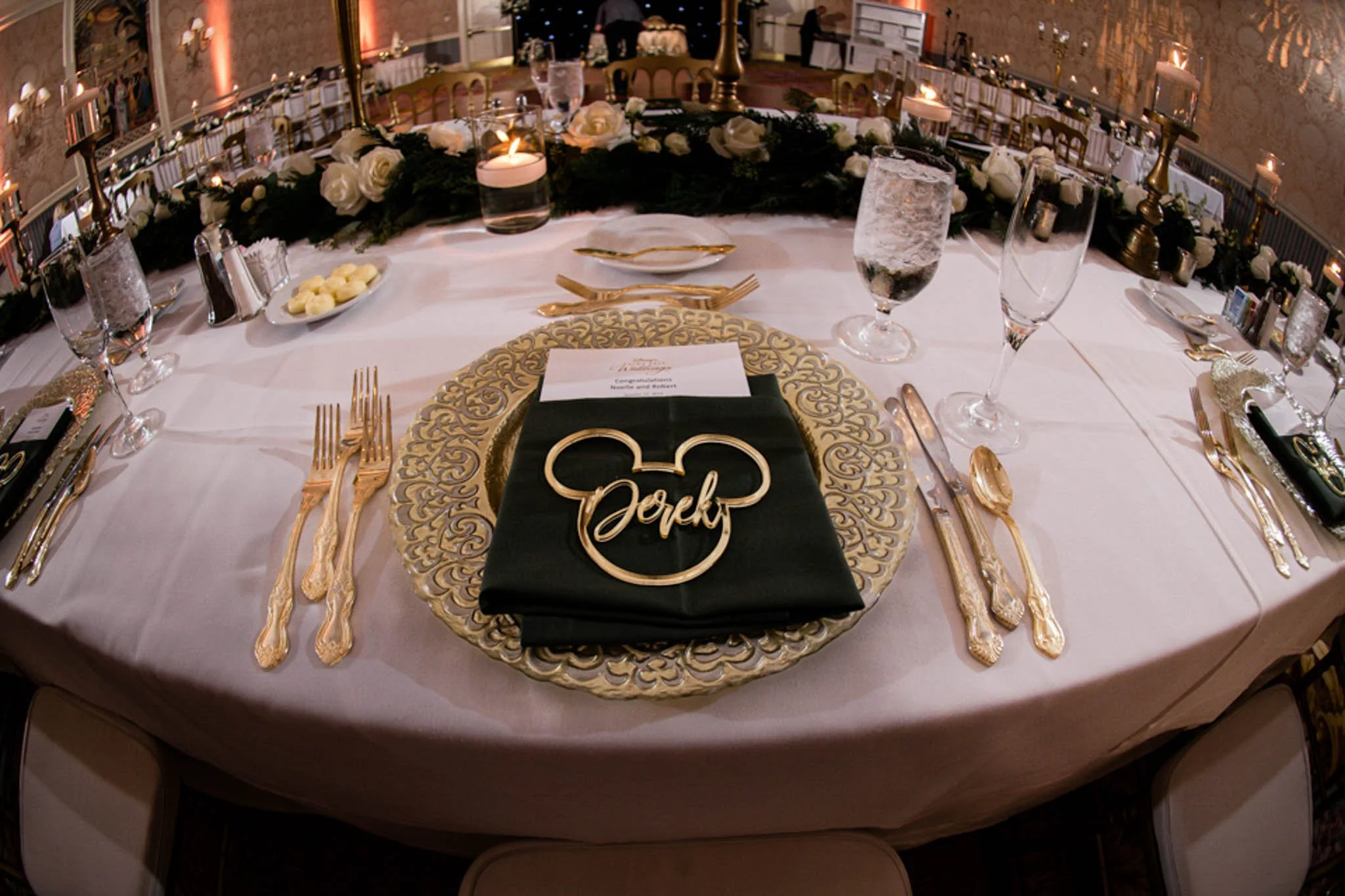 Elegant banquet table decorated with white tablecloth, gold cutlery, floral centerpiece, candles, and a black napkin with a gold Mickey Mouse head and the word 'Dork' in the center. Surrounding the table are chairs, with water glasses and place setti