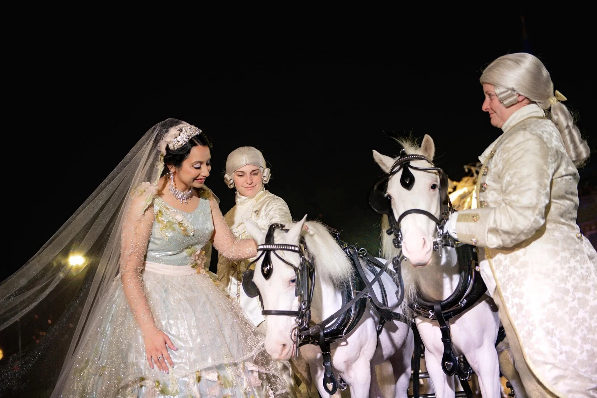 A bride in an elegant wedding gown and two men dressed in historical attire riding white horses with black bridles at night.
