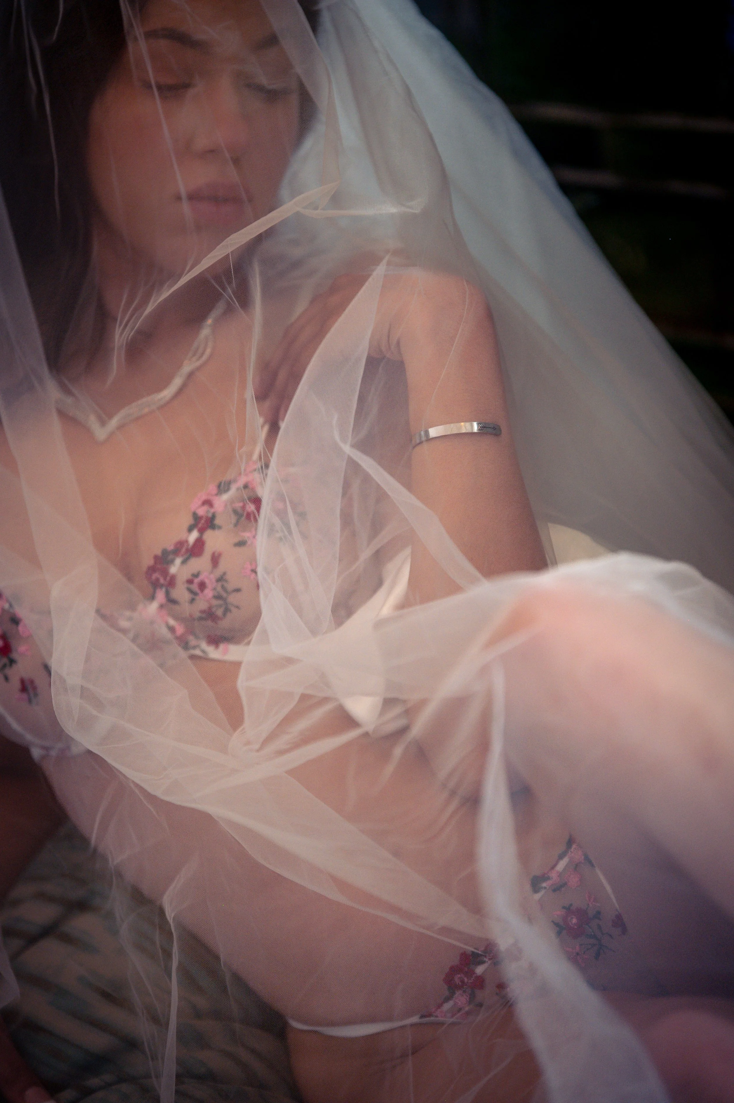 A woman wearing a wedding veil and floral lingerie, sitting with her eyes closed, partially covered by sheer fabric.