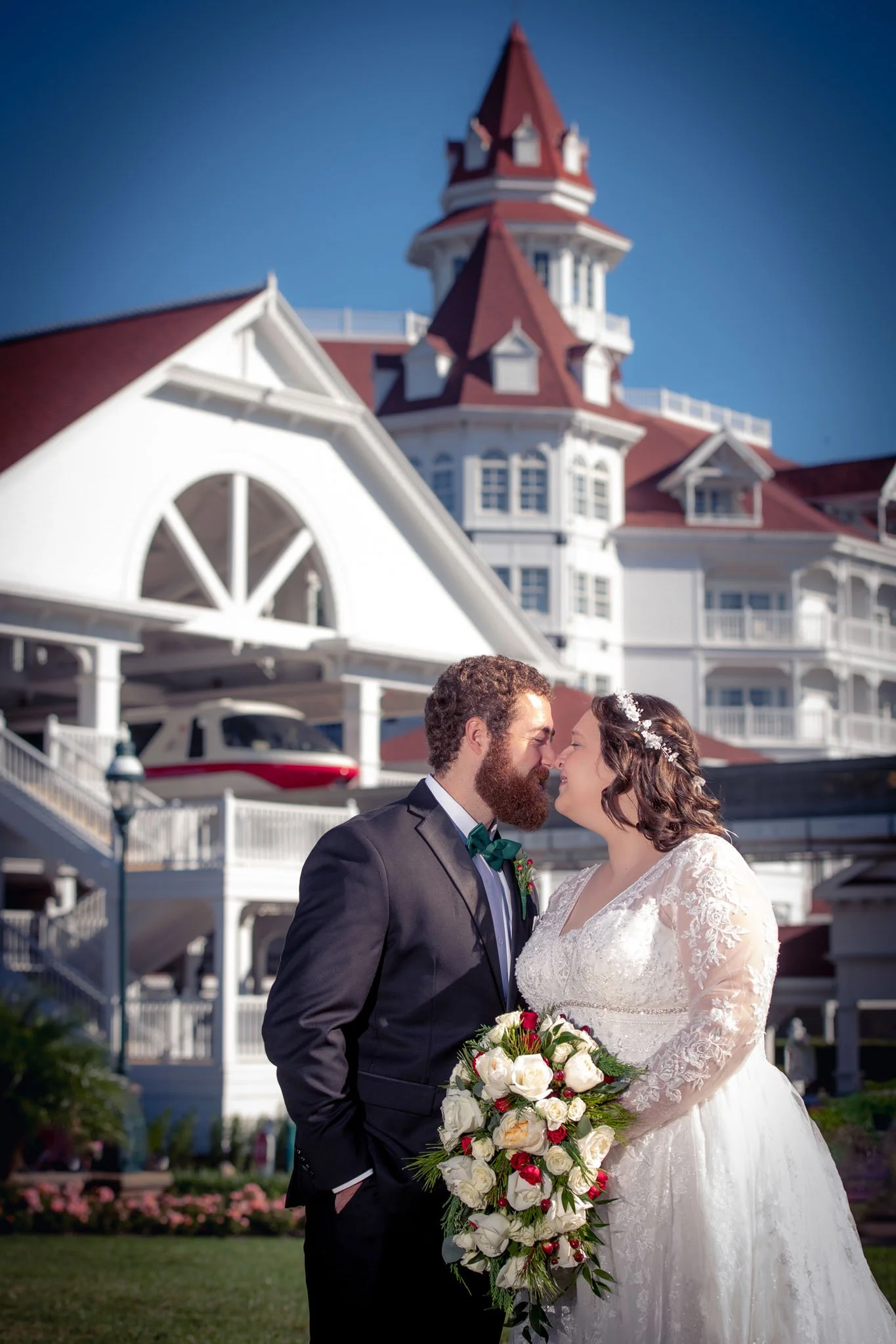 A bride and groom stand close together outdoors, about to kiss, with a large Victorian-style hotel or building with a tower in the background. The bride is holding a large bouquet of white and red flowers, and both are dressed in formal wedding attir
