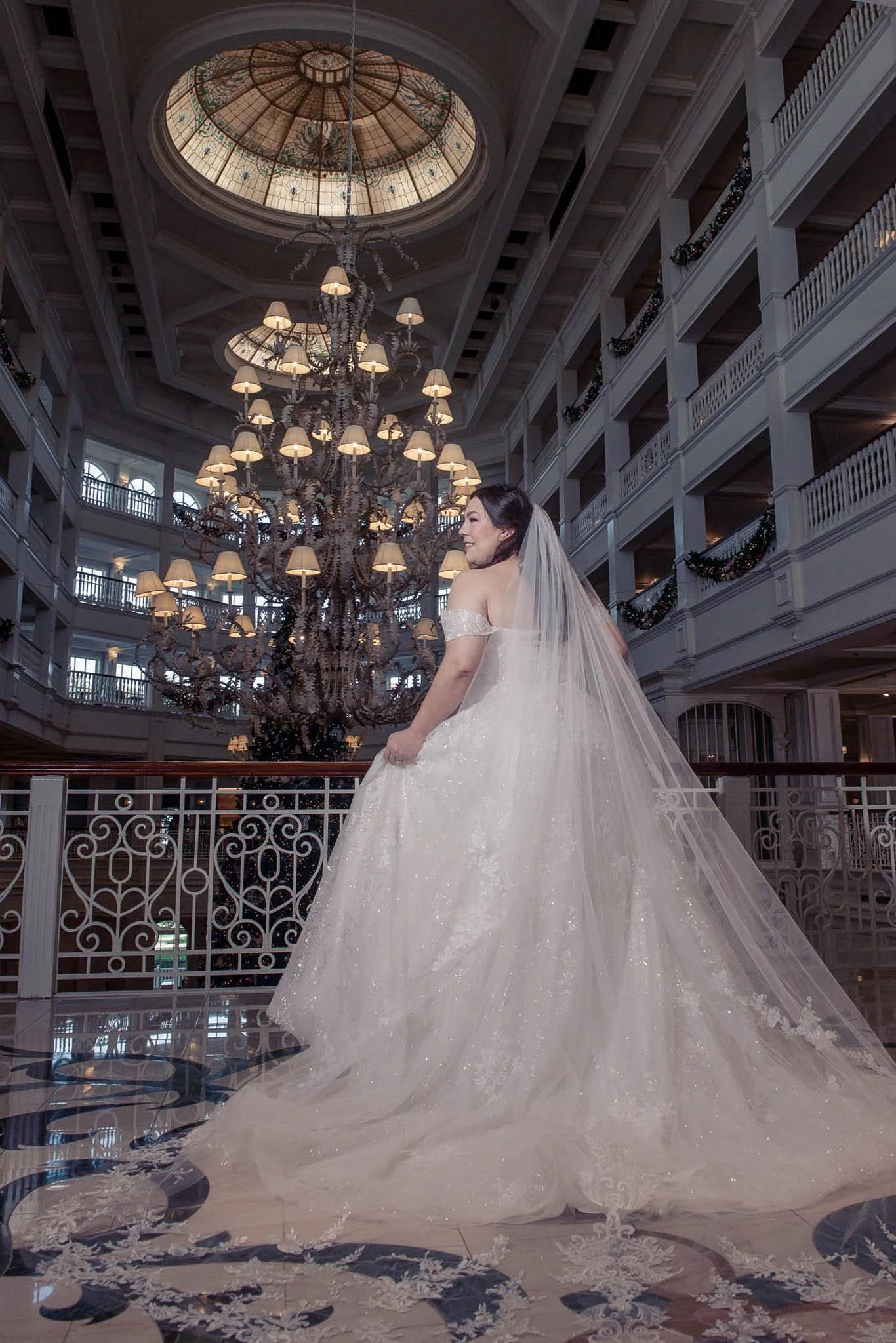 A bride in a white wedding gown and veil standing in a grand hotel lobby with multiple balconies, ornate railings, and a large chandelier, facing sideways with a smile.