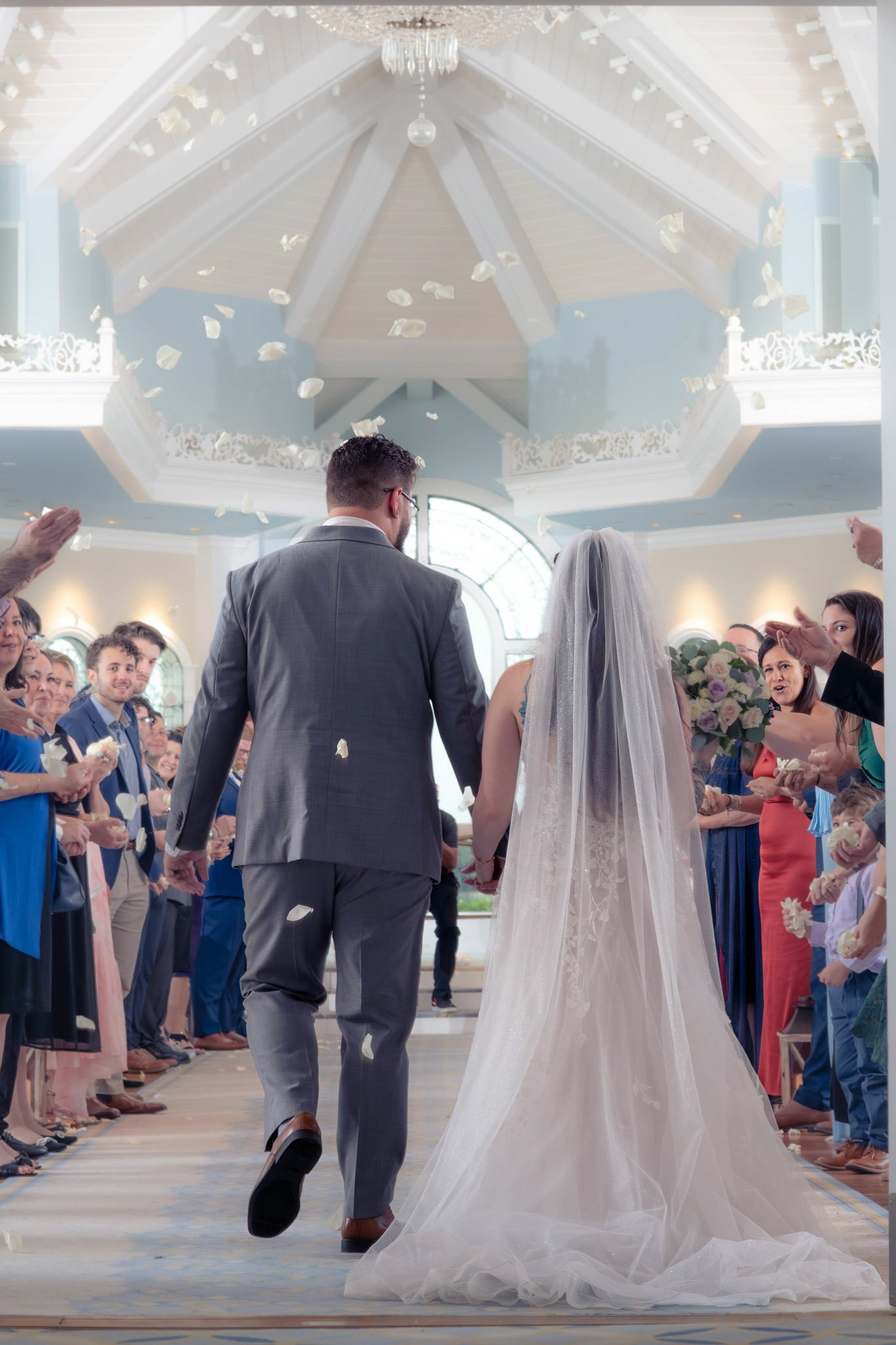 A wedding ceremony with a groom and bride walking down the aisle inside Disney's Wedding Pavilion, surrounded by guests and white rose petals in the air.