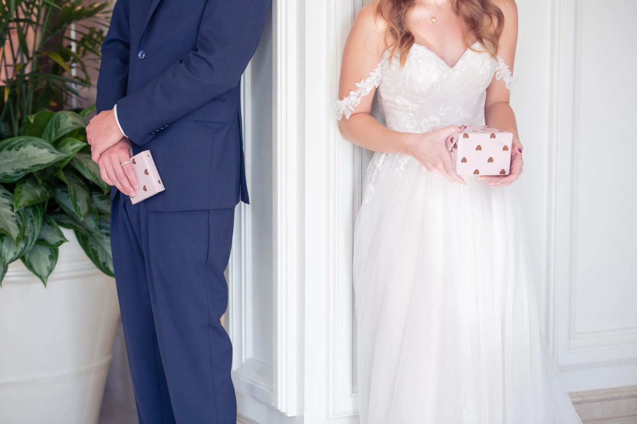 A bride and groom standing with their backs against each other, holding small gift boxes with heart patterns.