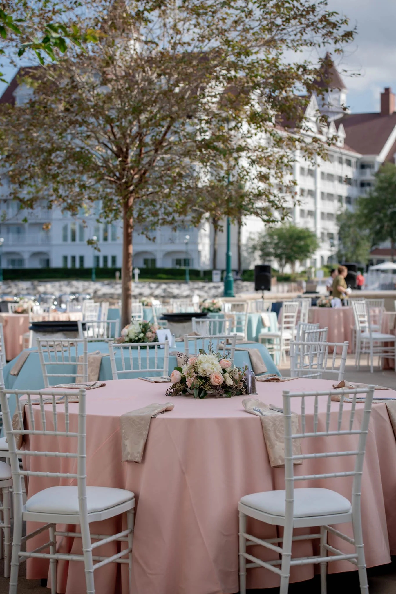 Outdoor wedding reception setup at The Grand Floridian with round tables covered in pastel pink and blue tablecloths, decorated with floral centerpieces, white chairs, and a tree in the background.