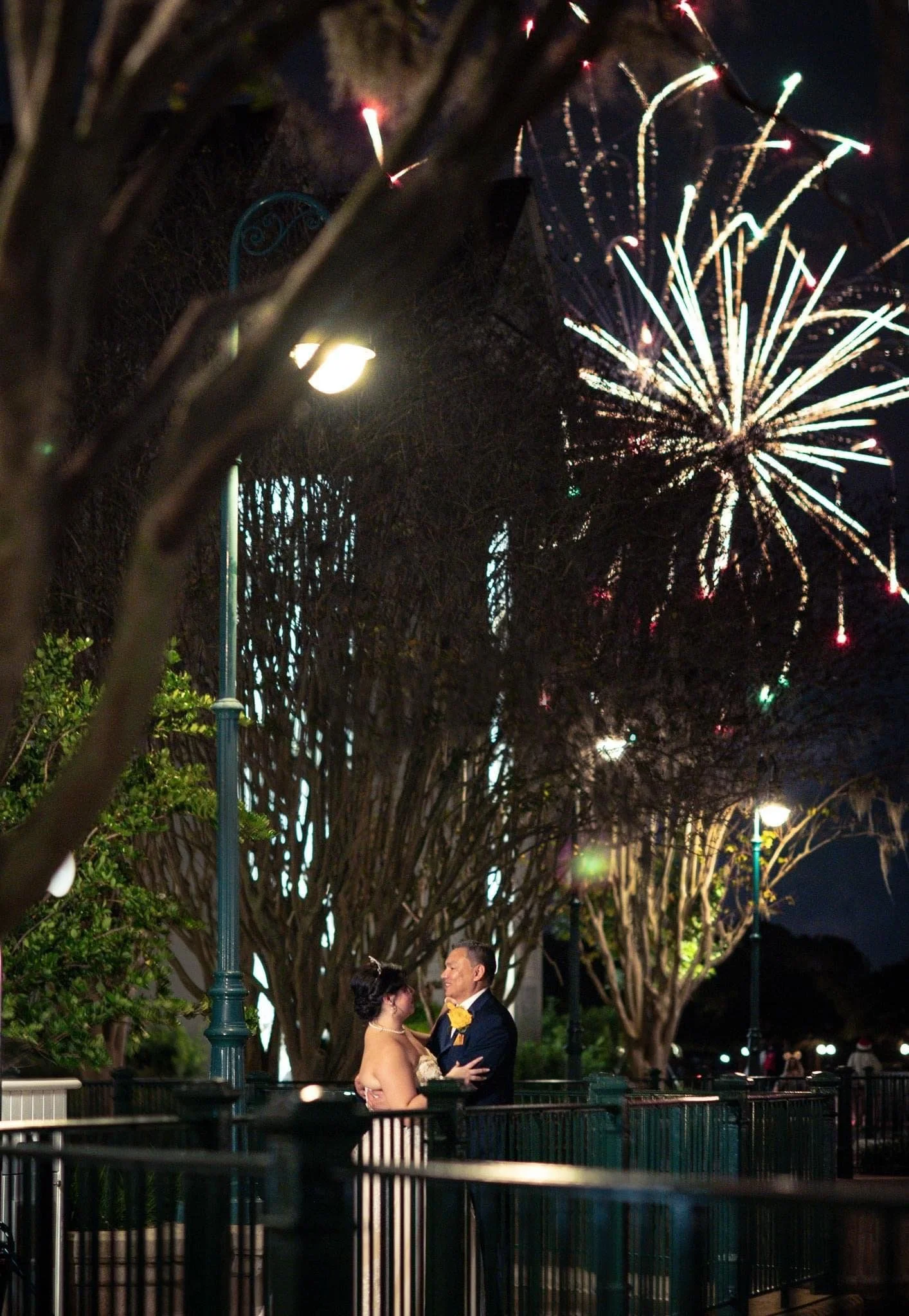 A couple sharing an intimate moment at night near a park railing, with Magic Kingdom fireworks lighting up the dark sky in the background.