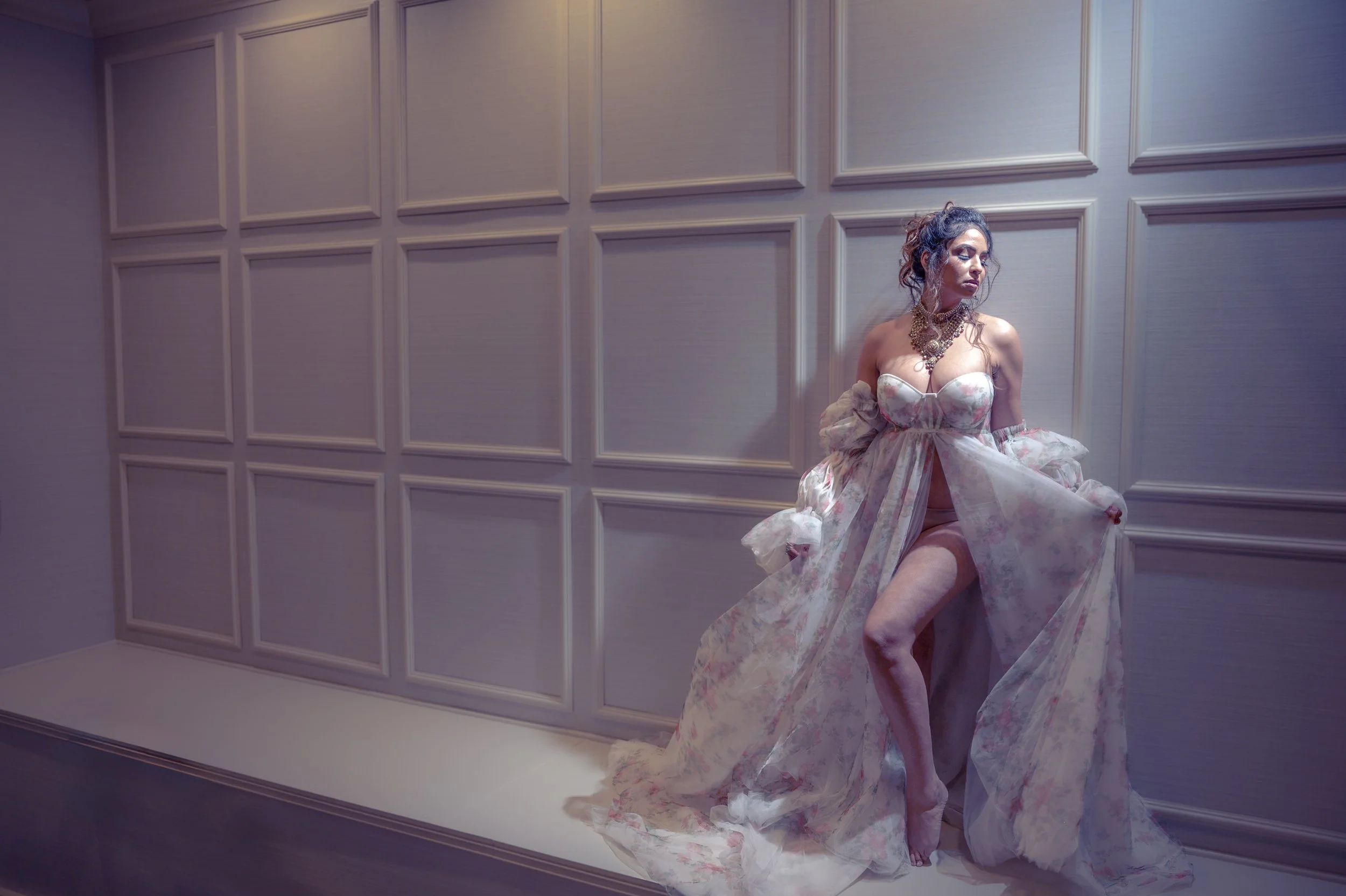 A woman in a floral strapless gown with a high slit, standing against a paneled wall, illuminated by soft lighting.