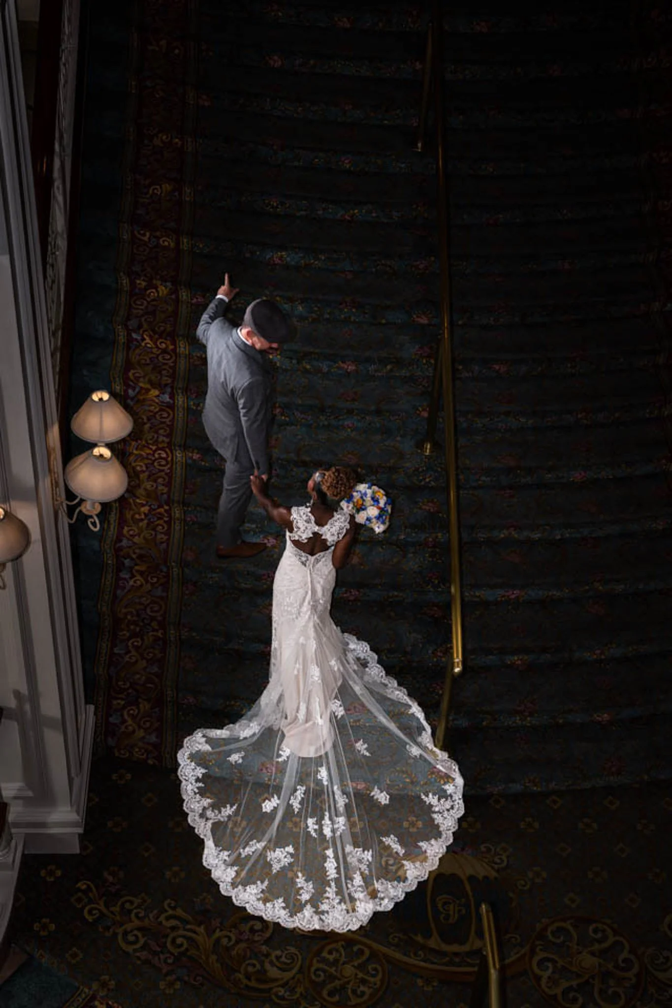 An overhead view of a man in a gray suit holding hands with a bride in a white wedding dress with a long train and a bouquet of flowers, walking on a dark ornate carpeted staircase.